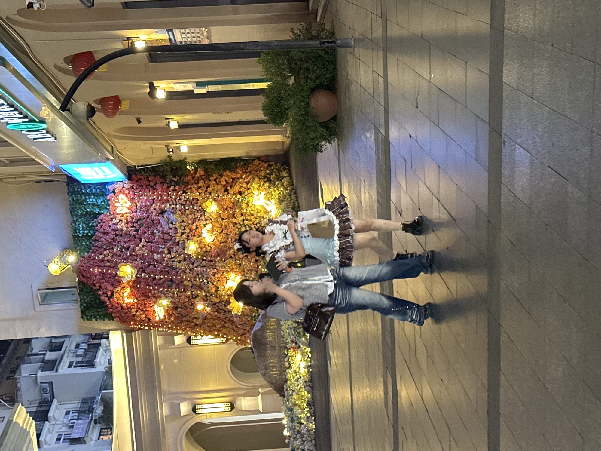 Two young women walk on a paved outdoor walkway at night. The woman on the left wears a checkered shirt and blue jeans, carrying a dark handbag. The woman on the right is dressed in a maid costume with a blue top and a ruffled brown skirt, holding a tablet and a shopping bag. Behind them, a wall is decorated with a dense arrangement of colorful flowers and bright lights, creating a festive backdrop. The surrounding architecture suggests a commercial or entertainment area, with illuminated storefronts and decorative lights. The scene is located in the Chongqing High-tech Zone, China.