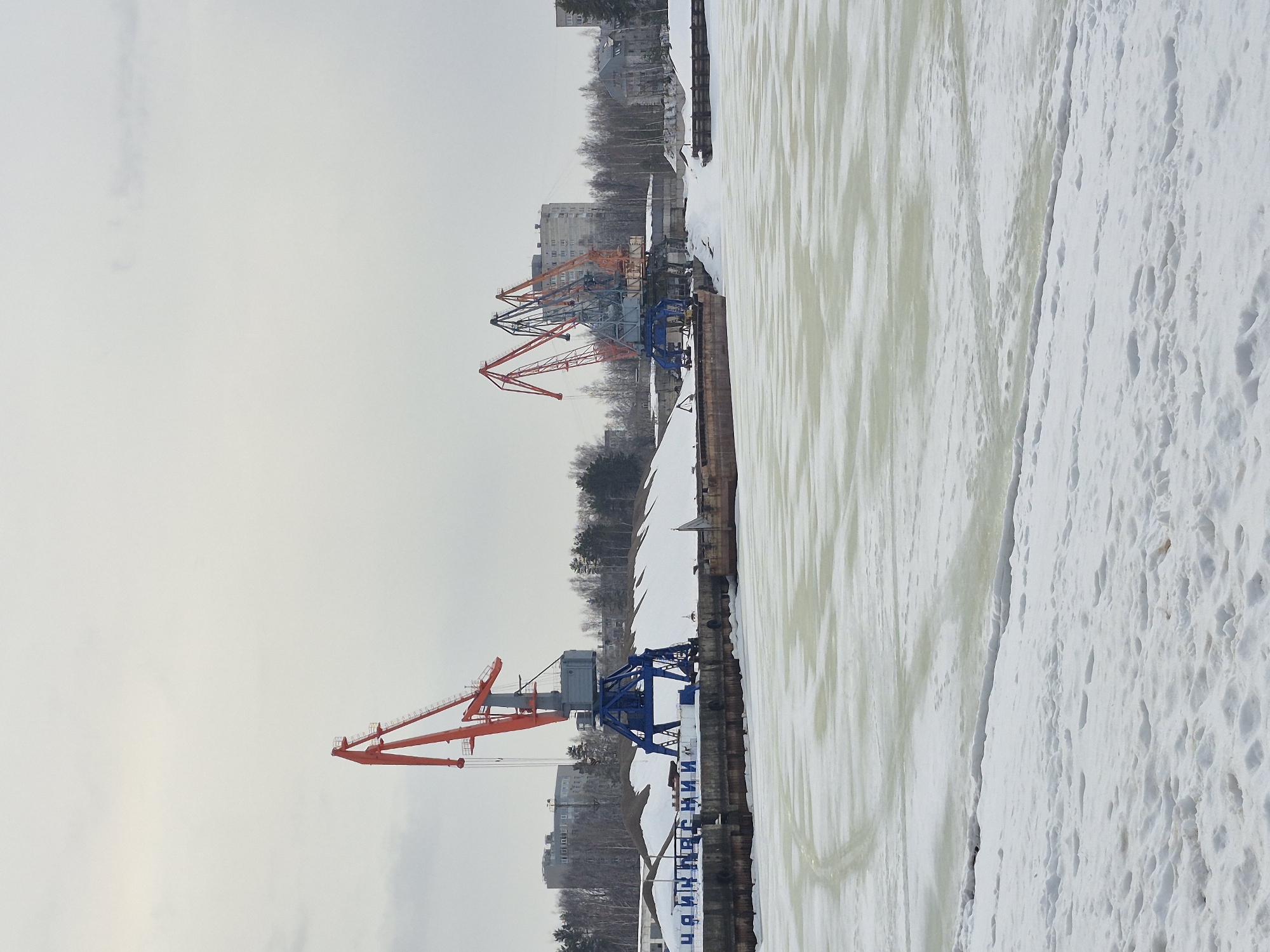 This image captures a winter scene at the port in Chaikovsky, Russia, likely situated on the Kama River or Votkinsk Reservoir, under a uniformly overcast sky.
The foreground presents an expanse of snow-covered ground marked with numerous footprints. Immediately beyond, a large body of water is frozen solid, displaying areas of yellowish-greenish ice and distinct linear patterns or tracks on its surface.
Along the far bank of the frozen water, a concrete dock wall runs horizontally. Blue Cyrillic letters, spelling "ЧАЙКОВСКИЙ" (Chaikovsky), are visible on this wall towards the left side of the frame. Behind the dock wall, substantial snowdrifts are accumulated. Three large gantry cranes dominate the middle ground: one on the left features an orange boom atop a blue support frame and a grey counterweight, while two additional cranes are clustered further right. One of these also has an orange boom and a blue base, with another, darker-colored crane with an orange jib positioned partially behind it. Flat-bottomed barges or similar vessels are docked along the wall, partially obscured by the snow and dock infrastructure.
The background consists of a dense, bare tree line, primarily deciduous, serving as a buffer between the industrial port and residential areas. Several multi-story apartment buildings are visible through and above these trees, forming part of the city's skyline. No individuals are present in the scene, and no specific actions or interactions are depicted.