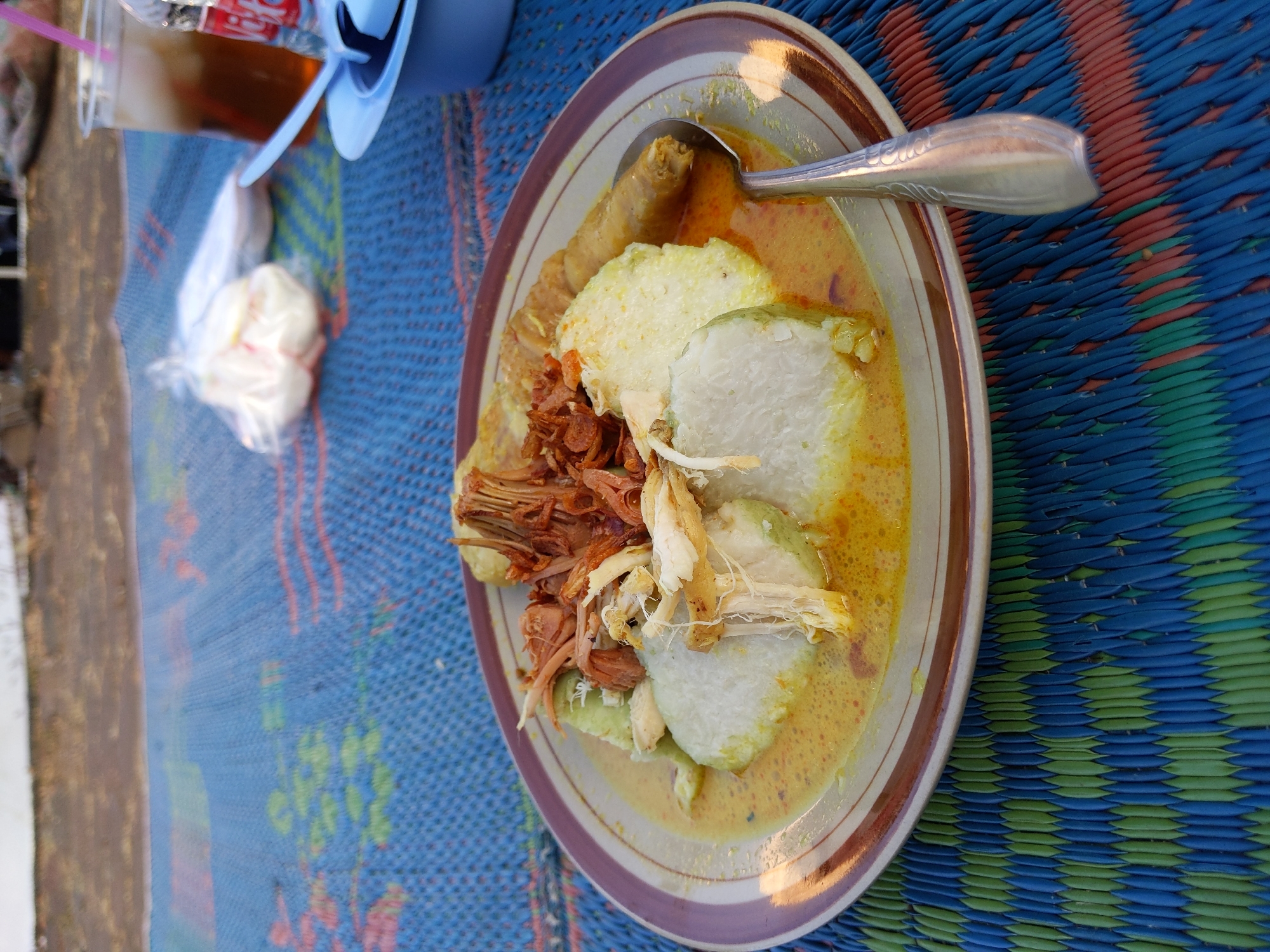 A close-up, eye-level, outdoor shot captures a plate of Indonesian food, possibly Lontong Cap Go Meh, served on a woven mat with a blue and green pattern. The meal consists of compressed rice cakes (lontong) submerged in a yellow curry broth, topped with shredded chicken and crispy fried onions. A piece of fried egg roll or similar item rests on the right side of the plate. A silver spoon is placed in the curry. In the background, a plastic cup with iced tea and a straw is visible, along with a plastic bag containing white items. The ground in the far background appears to be dirt. The scene is set in Nyamok, Indonesia.