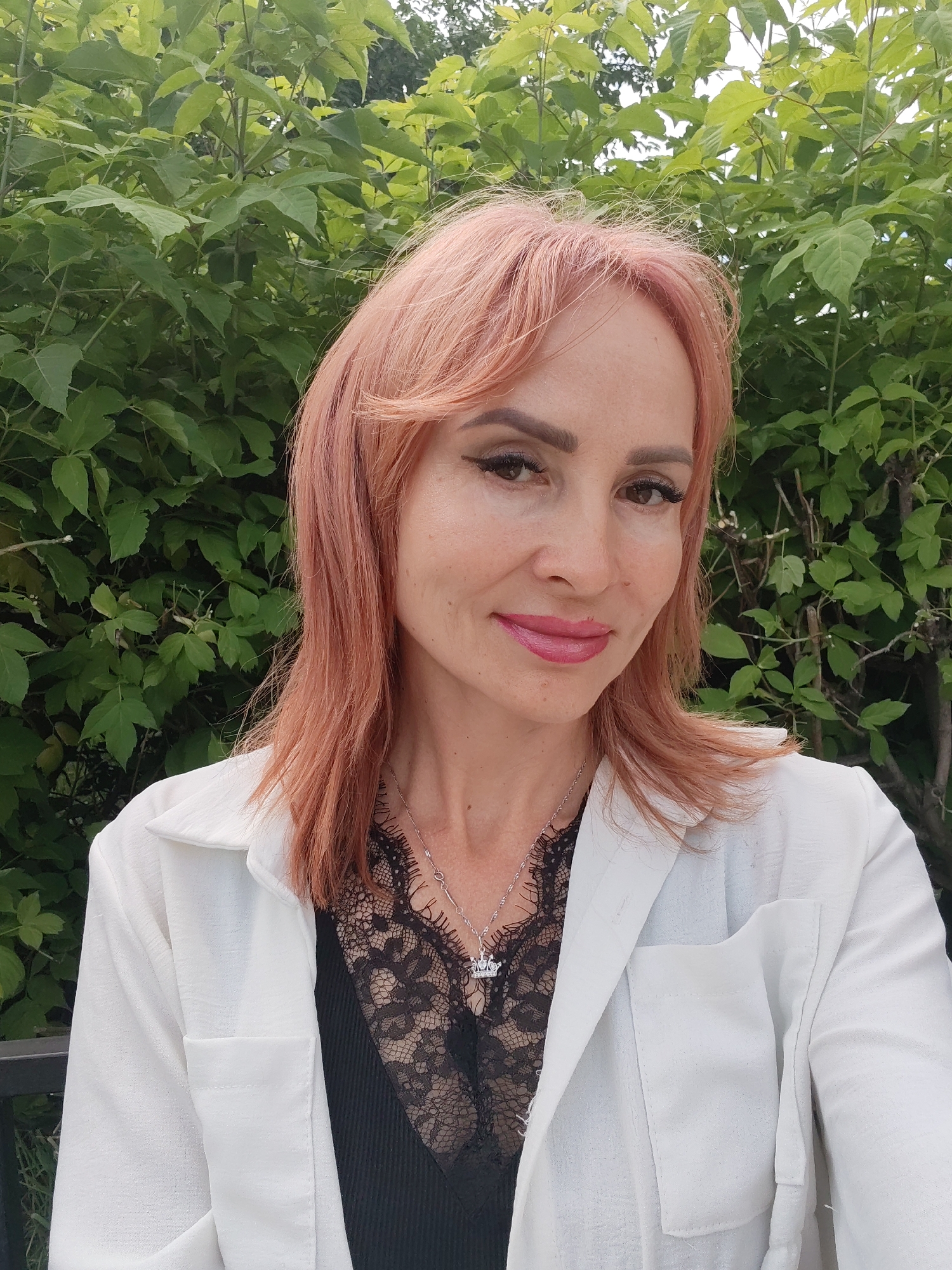 A medium shot shows a woman with pink hair and makeup posing outdoors in front of a green leafy background. She is wearing a white jacket over a black lace top and a silver necklace with a crown pendant. Her hair is styled with bangs and is slightly messy. The woman's eyes are looking directly at the camera, and she has a slight smile. The background is filled with lush green foliage, suggesting an outdoor, natural setting. The image appears to be a selfie taken during the day. No specific location in Chaikovsky, Russia, can be identified from the image.