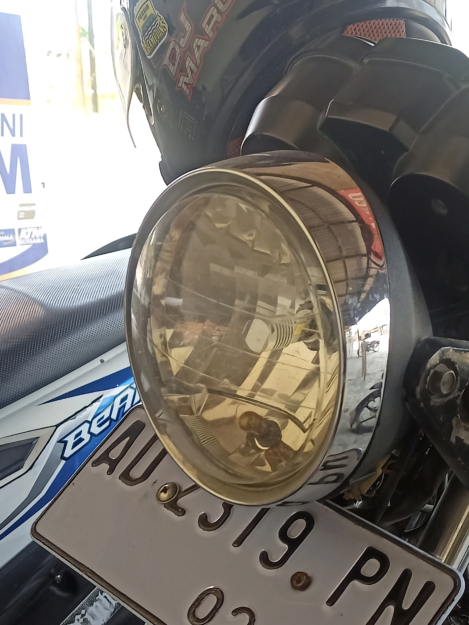 A close-up shot captures the front headlight and license plate of a motorcycle, with a helmet visible in the upper portion of the frame. The motorcycle's headlight has a chrome rim and a yellow-tinted lens. A portion of the motorcycle's seat, textured with a carbon fiber pattern, is visible to the left of the headlight. A white license plate with dark lettering, reading "AU 40 519 PN," is mounted below the headlight. The background is out of focus but appears to be an indoor or covered outdoor area with sunlight filtering through, suggesting a location like a garage or a covered parking spot within a city like Gabungan, Indonesia. The helmet on top has stickers, one of which appears to be for "PERSID 1953" and another with "DJ MARCO."