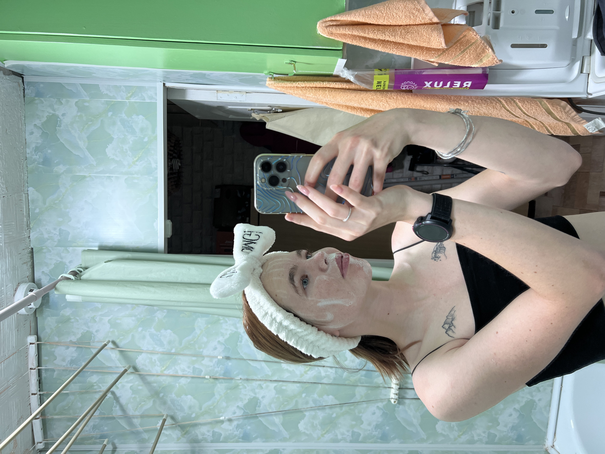 The image captures a young woman with fair skin and light brown hair tied in a ponytail, wearing a white fuzzy headband with bunny ears. She has a white facial mask applied to her face. She is wearing a black spaghetti strap top and a black smartwatch on her left wrist. She is holding a smartphone in her right hand, appearing to take a selfie in a bathroom. The bathroom walls are tiled with a green and white marbled pattern. In the background, a green curtain hangs, and a clothes drying rack is visible. To the right, orange towels are hanging, and a purple bottle with text is also visible. A tattoo of mountains and a wave is visible on her chest. The scene is likely indoors, in a residential bathroom, possibly located in the city of Chaikovsky, Russia, given the context provided.