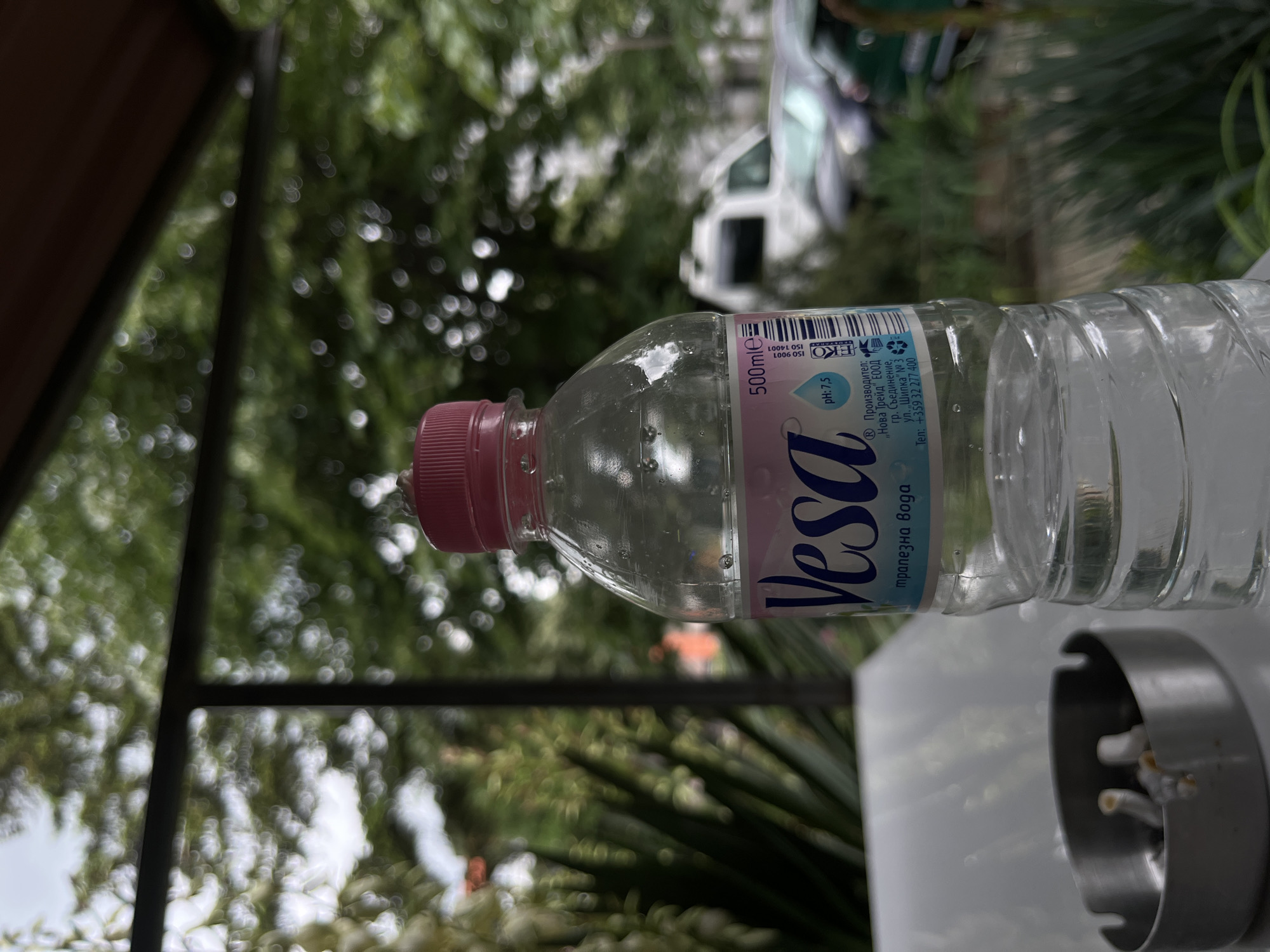 The image displays an outdoor scene, likely a patio or garden area, during daylight. A clear plastic water bottle with a pink screw cap is the prominent subject, resting horizontally on a light-colored surface, presumably a table. The bottle contains water with visible air bubbles.
The label on the bottle is largely in Bulgarian, identifying the brand as "Vesa" (Веса) and the contents as "трапезна вода" (table water). Notable details on the label include "500mle" and a "pH: 7,5". The manufacturer is listed as "„Нова Трейд" ЕООД" from "гр. Съединение, ул. „Шипка" № 3", with a phone number "+359 32 277 400". Certifications like ISO 9001, ISO 14001, an "EKO" logo, and a recycling symbol are also visible.
To the right of the bottle, on the same surface, a grey cylindrical ashtray containing several white cigarette butts is present. The background is softly blurred, featuring lush green foliage from trees or bushes, suggesting an natural outdoor setting. Dark structural elements, possibly part of a building or pergola, can be seen in the upper parts of the background. A faint, out-of-focus white vehicle is discernible through the distant greenery. The bright, natural lighting indicates it is daytime. The Bulgarian text on the bottle aligns with the provided location context of Shumen, Bulgaria, despite the manufacturer's listed location in Saedinenie. The scene evokes a casual outdoor moment.