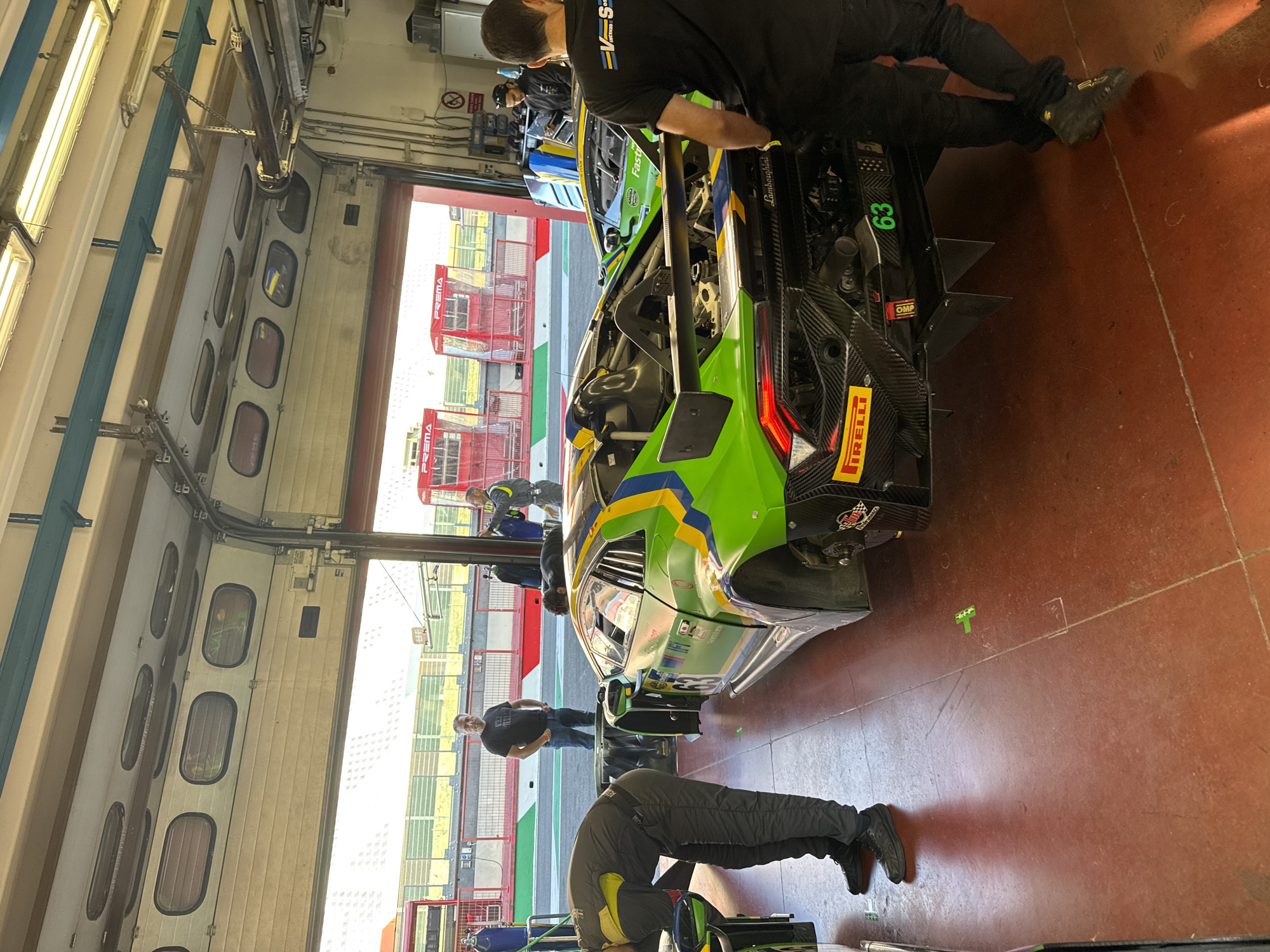 The image depicts a garage area at the Mugello Circuit in Scarperia, Italy, likely during a motorsport event. In the foreground, a green and yellow race car, possibly a Lamborghini, is parked. A person in a grey racing suit is bent over the car, appearing to work on it. Another person in a black t-shirt and dark pants stands to the right, facing away from the camera and looking at the race car. Further back, two other individuals are visible near the open garage doors. The garage doors themselves are elevated, revealing a track environment with tiered seating and red structures beyond. The floor of the garage is a reddish-brown color, and the walls are a light grey. There are various tools and equipment visible within the garage space. The overall scene suggests a pit stop or maintenance activity for the race car.