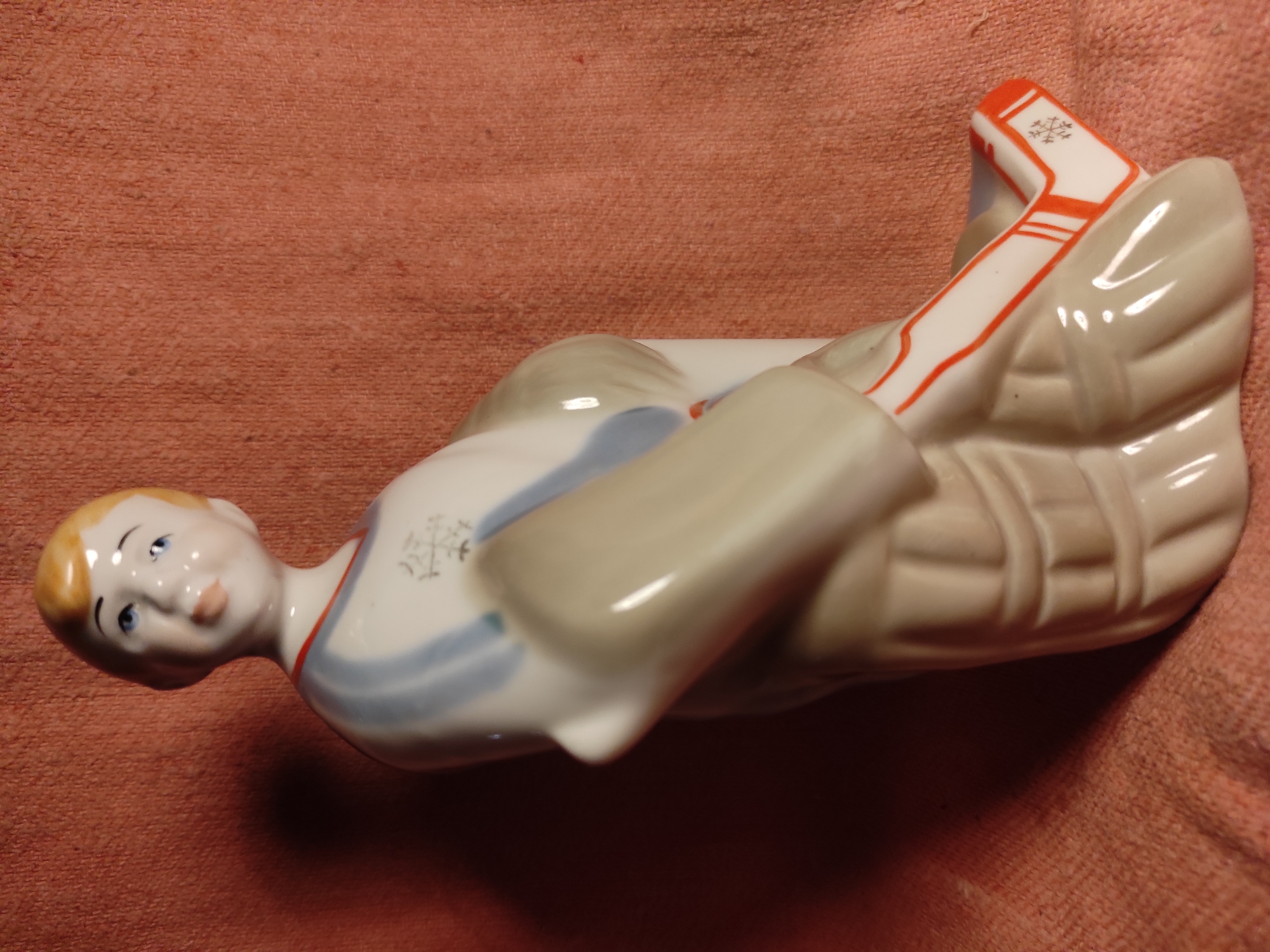 A light pink, textured background with a subtle weave pattern supports a porcelain figurine of a young boy. The figurine is predominantly white with accents of light grey, pale blue, and orange. The boy has blond hair, blue eyes, and rosy cheeks, with a slight smile on his lips. He is wearing a white outfit with light blue stripes across the chest and what appears to be protective leg gear detailed with orange lines and a small snowflake emblem on the right shin. His posture is a relaxed lean, with his right arm resting across his body and his left arm bent at the elbow. The figurine is positioned in a three-quarter view, with the boy's face turned slightly towards the viewer. The lighting casts a shadow to the left of the figurine. No information about the location in Chernihiv, Ukraine is discernible from the image itself.