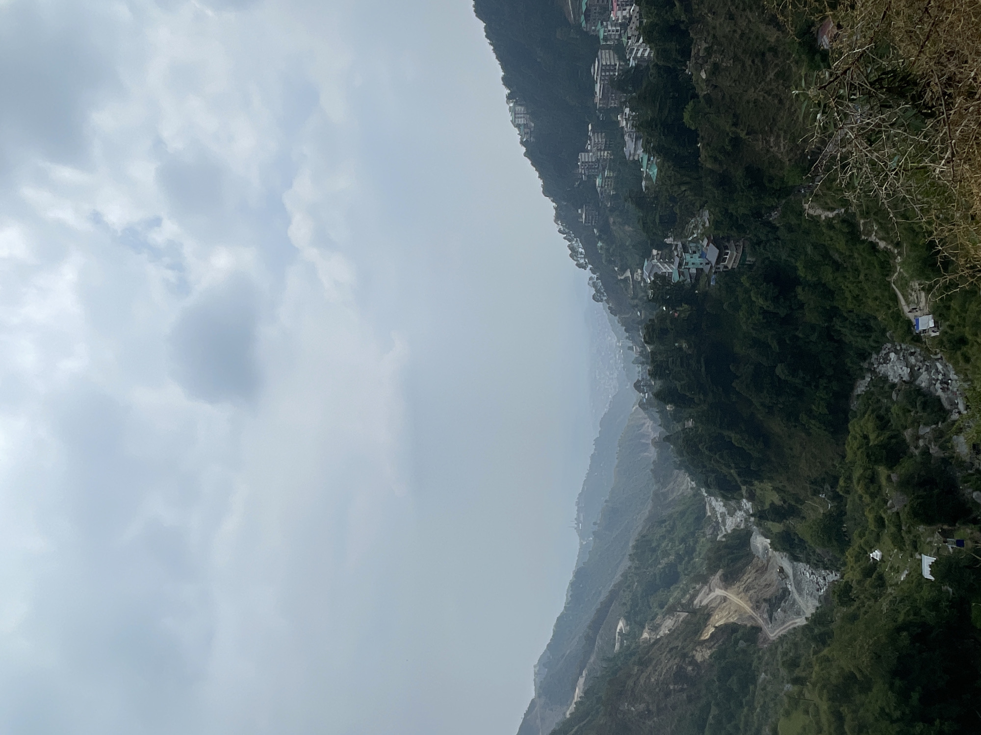 The image presents a landscape view in what appears to be the mountainous region of Bhagsu, India, under an overcast sky. The scene is dominated by steep, densely vegetated slopes of the Himalayas. There are buildings and structures nestled along the hillside, indicating residential or commercial development. A quarry-like area with exposed earth is visible further down the slope, hinting at construction or mining activity. The cloudy sky creates a muted, hazy atmosphere, obscuring distant details. Bare branches are seen on the right side of the frame.