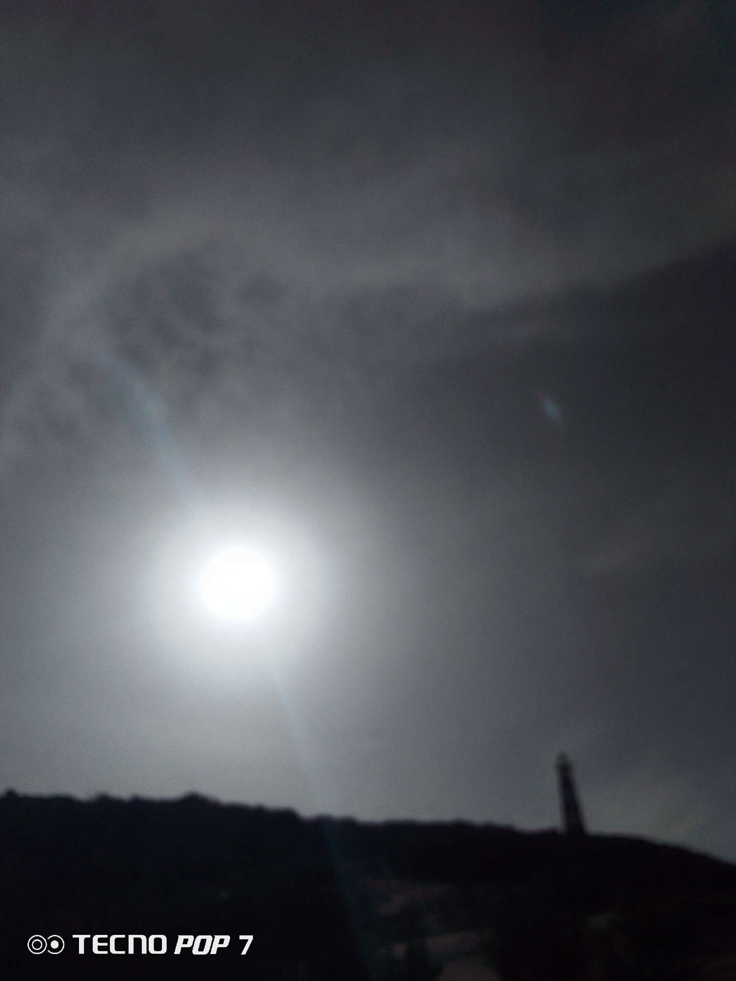 The image depicts a bright sun obscured by a gray, cloudy sky. In the foreground, a dark, silhouetted landmass is visible with a tall, indistinct structure resembling a lighthouse on top. The words "TECNO POP 7" are displayed in the lower left corner. The location is most likely Pedro González, Venezuela, indicated by the prompt. No people or interactions are visible in the image.