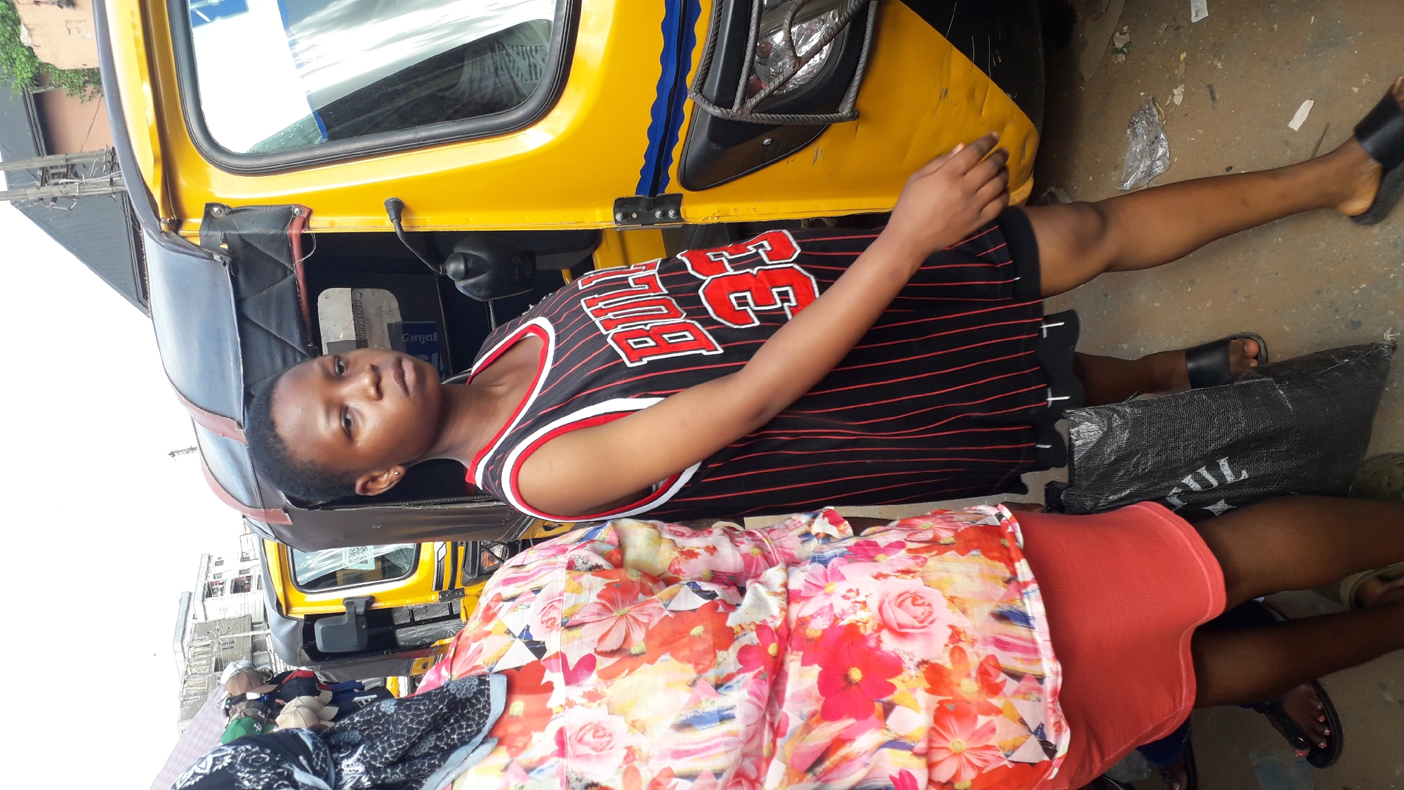 A woman in a Bulls jersey lies next to a yellow tuk-tuk in Onitsha, Nigeria. Another person in a floral top and orange shorts lies near her. The scene appears to be an outdoor street setting.