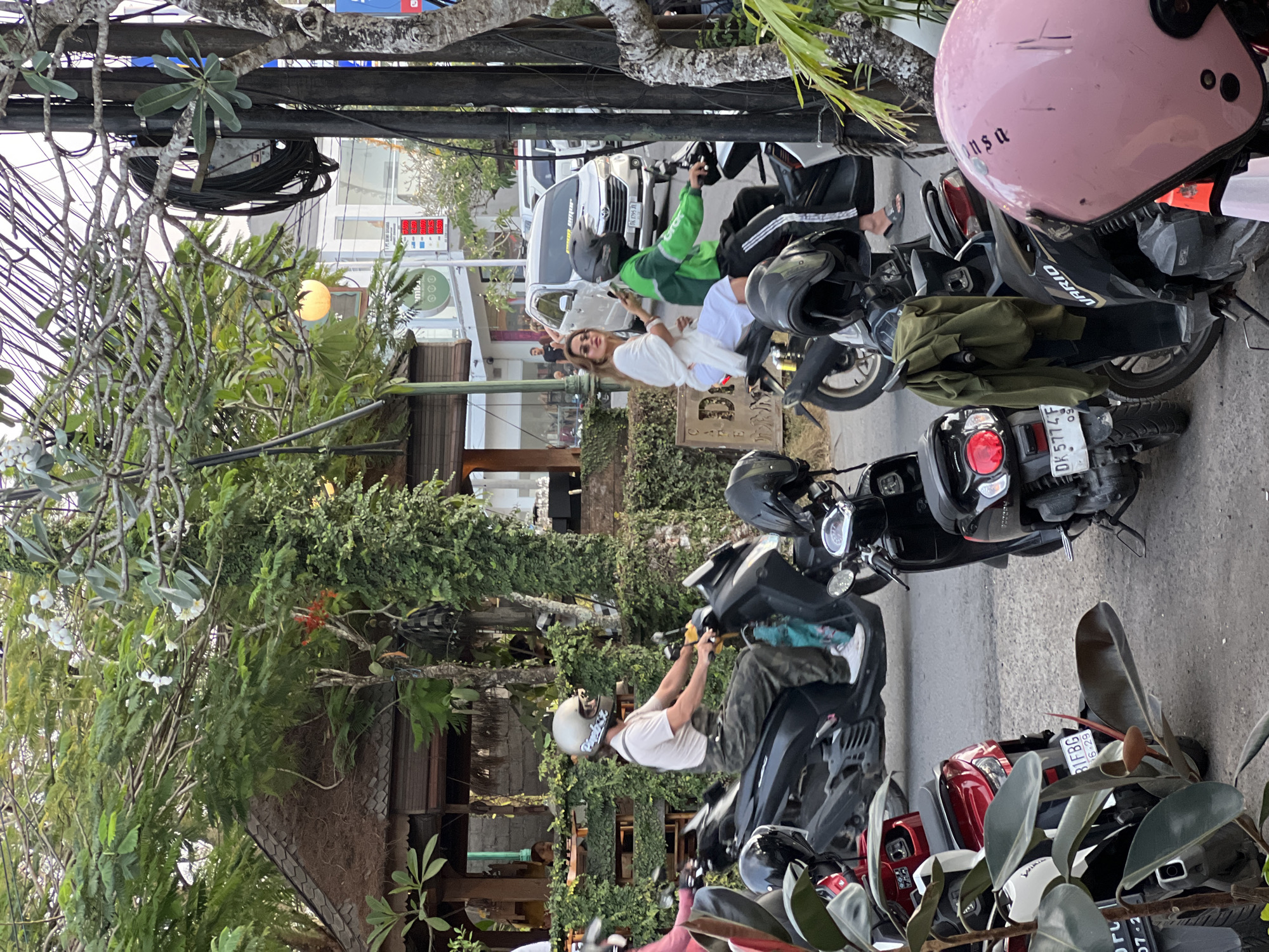 In Kuta Selatan, Indonesia, an outdoor scene depicts several parked motorcycles. In the foreground, a pink helmet sits on a motorcycle, with "ansa" written on it. To its left, a black motorcycle with a green garment hanging from its handlebars and "VARIO" visible on its side. Behind that, a black scooter with "DK 5774 F" license plate is parked. Further back, a man wearing overalls and a white t-shirt rides a black scooter. To his right, a woman in a white dress and a man in a green jacket ride a scooter. In the background, a white car is visible, along with buildings and lush greenery. The area appears to be a street, possibly near a restaurant or shop.