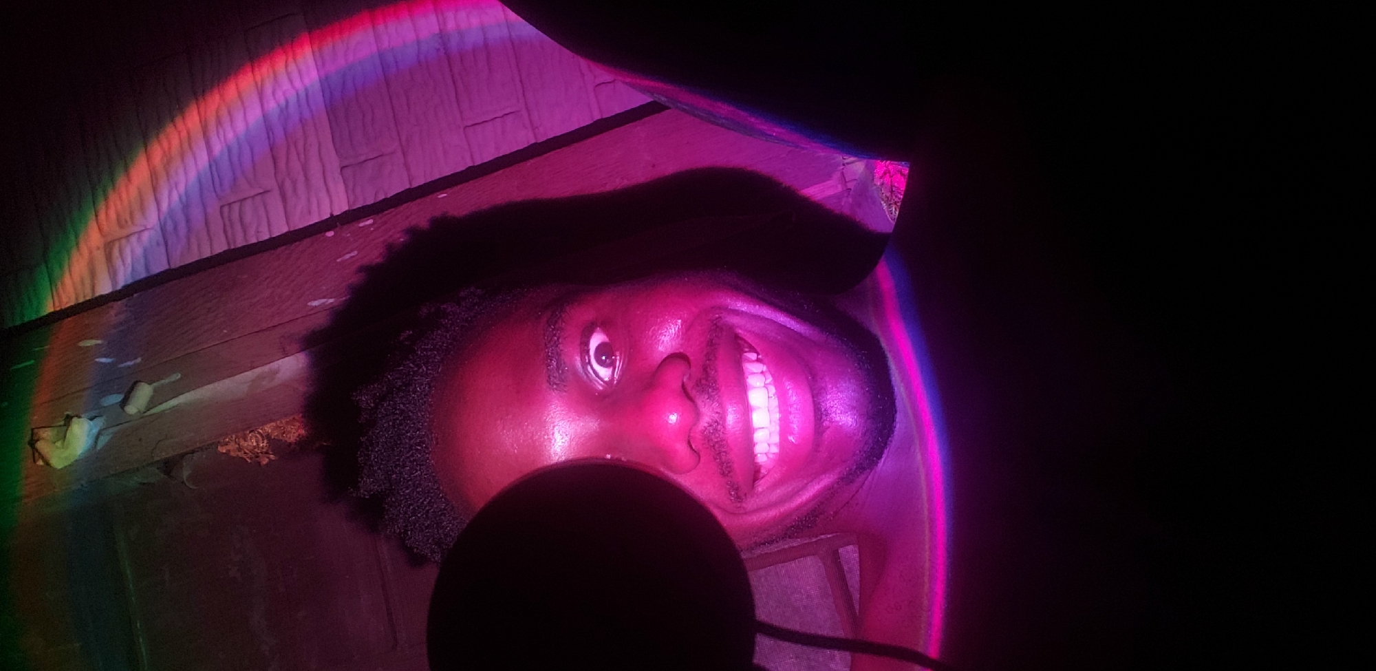 A close-up, low-angle selfie shows a Black man with dark, short hair, wide eyes, and a bright smile. The man is illuminated by a pinkish-purple light, with a rainbow-colored light projected across the background. He appears to be in a dimly lit, indoor space with wooden planks visible above his head and a brick-patterned wall to his right. A large, dark, circular object, possibly a microphone or camera lens, is positioned to the left of his face, partially obscuring it. The overall impression is a playful and artistic self-portrait. The location is identified as Narayi, Nigeria.