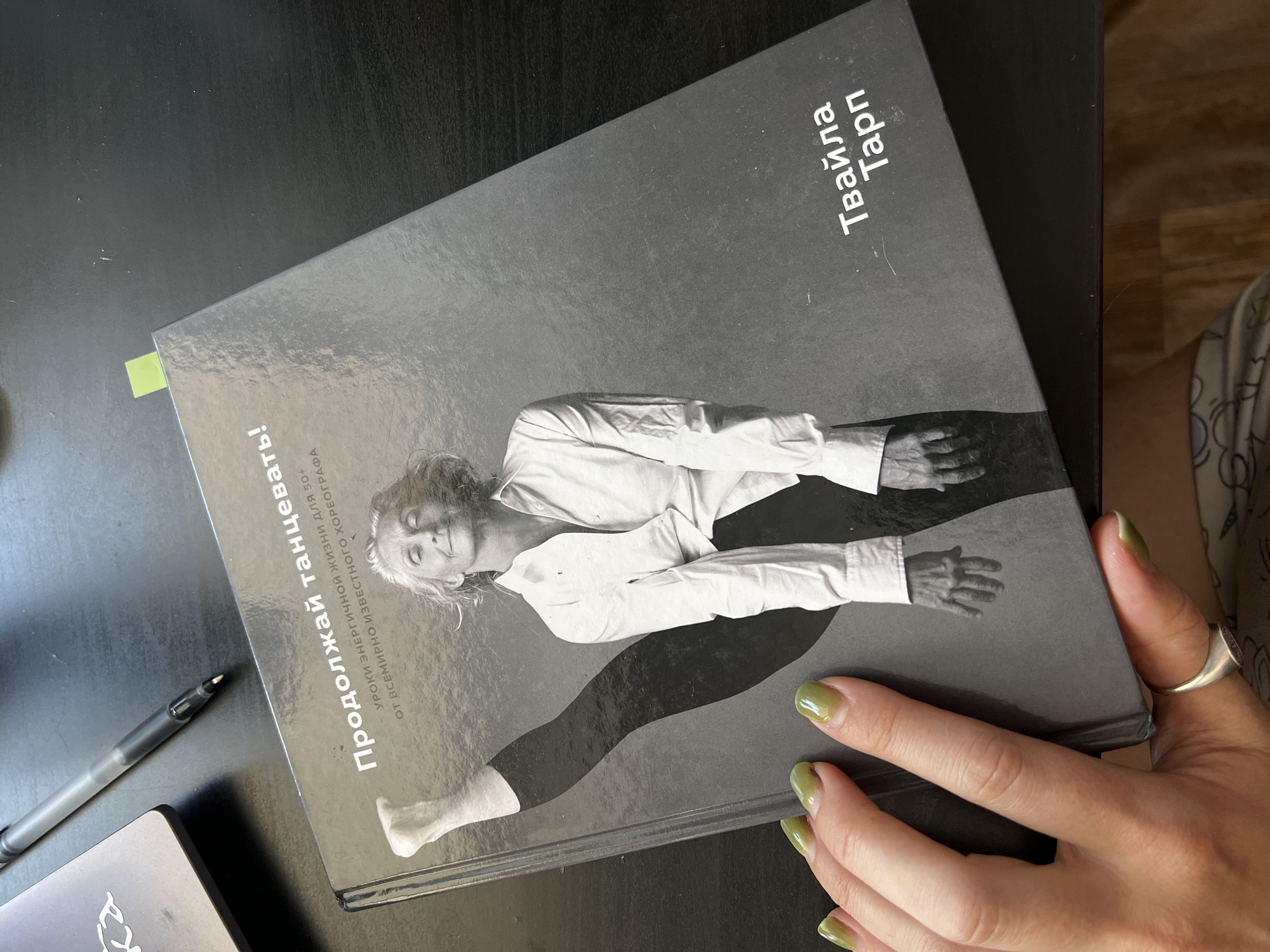 A black hardcover book titled "Continue Dancing!" with a subtitle about energetic lessons for those over 50 from a world-renowned choreographer is placed on a dark, reflective surface. The book's cover features a black and white photograph of an older woman in a white shirt, with one leg raised high. A woman's hand with green nail polish is holding the book on the left side of the frame, with a silver ring on her thumb. A black pen lies on the surface to the upper left of the book, next to a partially visible laptop. The scene is likely indoors, on a desk. There is no discernible information within the image or its context to indicate a specific location in Novosibirsk, Russia.