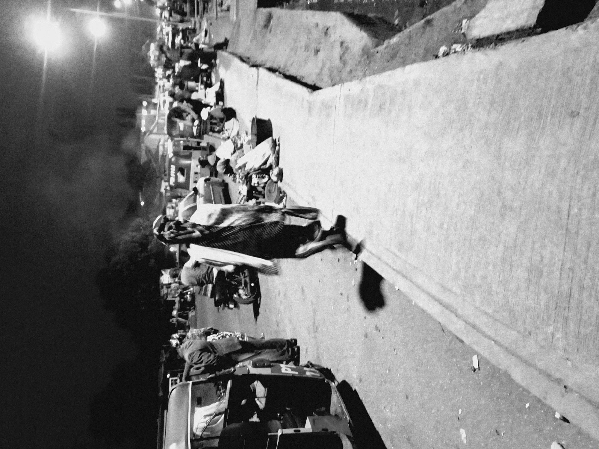This black and white photograph captures a bustling nighttime street scene in Bukuru, Nigeria, filled with people, vehicles, and market activity. The image exhibits strong contrasts and significant grain, characteristic of low-light photography.
In the foreground, a concrete barrier or low wall extends from the bottom right, separating a paved ground area from a higher, possibly dirt, embankment. Scattered debris is visible on the paved surface. To the bottom left, the front section of an auto-rickshaw or similar compact public transport vehicle is partially visible, with the letters "P I" faintly discernible on its side. Another individual is seated inside this vehicle.
The midground is densely populated with people, suggesting a vibrant market or communal gathering. Many individuals appear to be vendors or shoppers, moving among various makeshift stalls or structures. A prominent figure, centrally located, is seen carrying a large, dark bundle on their head or shoulder, walking away from the camera. A motorcycle is parked further back among the crowd. A small, dark animal, possibly a dog, can be seen near the concrete wall in the middle ground.
The background is dominated by the dark night sky, punctuated by several intensely bright, artificial light sources that cast strong glare and shadows across the scene. These lights illuminate what appears to be smoke or steam rising from the crowd. The presence of numerous people and improvised selling points indicates a lively commercial area operating after dusk. The atmosphere is active and typical of an evening marketplace in an urban or semi-urban setting.