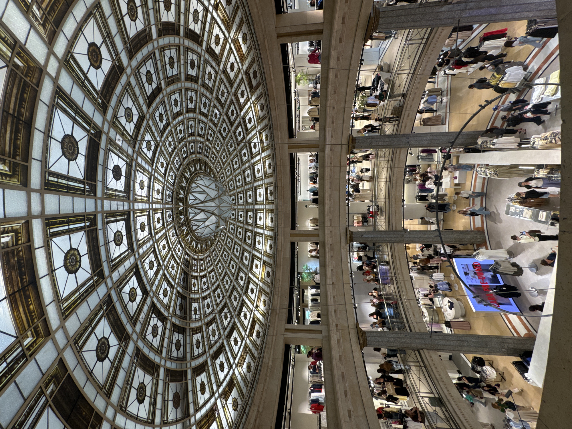 The image captures an interior view within a retail establishment, specifically Zara in Barcelona, Spain. The focal point is an ornate, domed ceiling adorned with intricate geometric patterns and decorative floral motifs. Sunlight streams through the glass panes of the dome. Below, the multi-level retail space bustles with activity. Shoppers browse clothing displays on various floors, visible through openings and balconies. The architecture features stone columns and railings that provide vertical structure to the space. People are discernible engaging in shopping activities, such as examining clothing racks. The environment is well-lit and suggests a sense of grandeur.