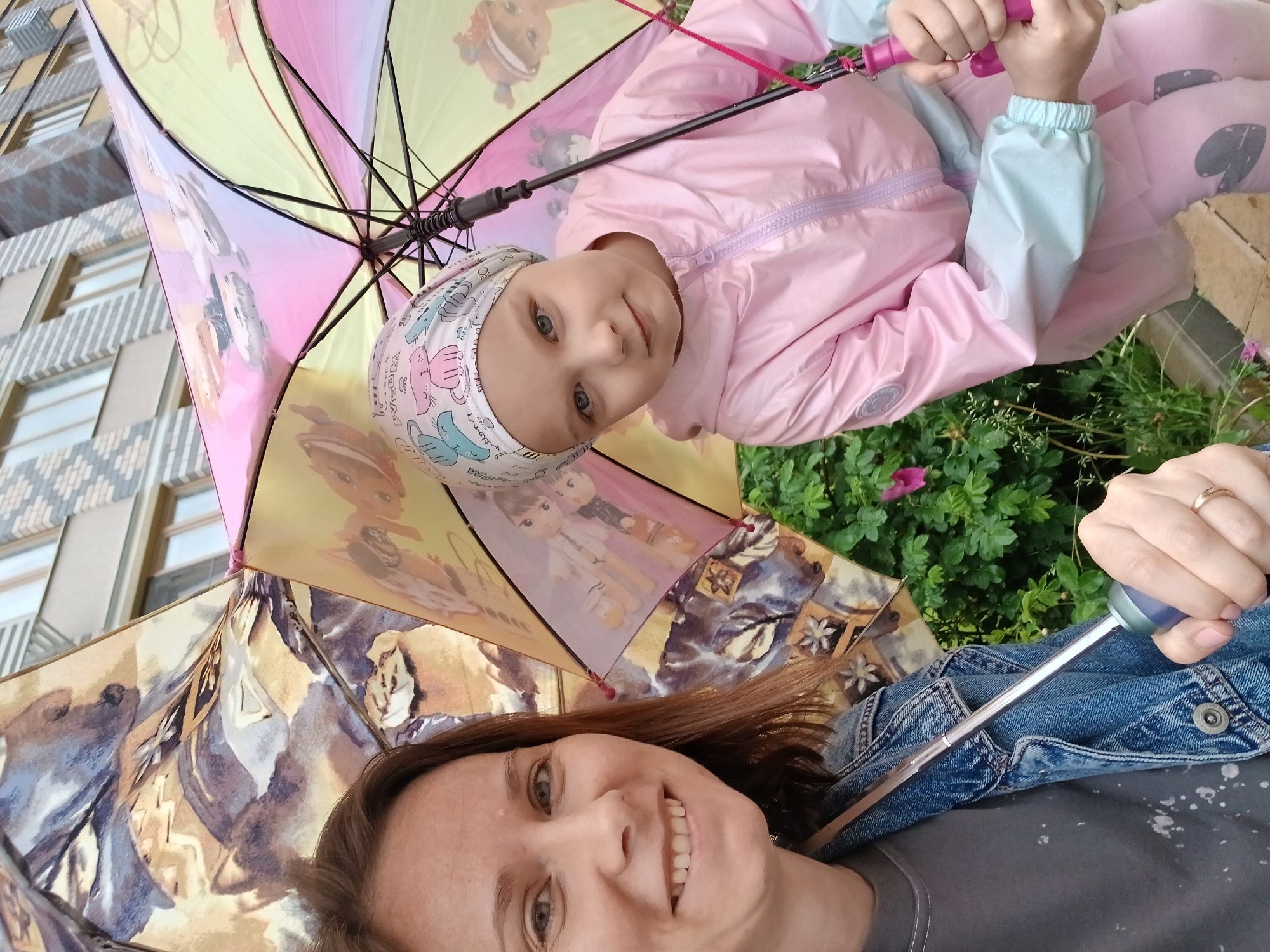 The image shows a woman and a young girl sharing an umbrella outdoors. The woman, in the foreground on the left, is smiling and wearing a denim jacket over a grey t-shirt. She is holding the handle of an umbrella. The girl, on the right, is wearing a pink hooded jacket and pink leggings. She is also holding onto the umbrella, which is decorated with cartoon characters and has a pink, yellow, and purple color scheme. The background features a building with a brick facade and windows, and green foliage with a pink flower. The setting appears to be a residential area, possibly a courtyard or street in Yekaterinburg, Russia, given the architectural style of the building.