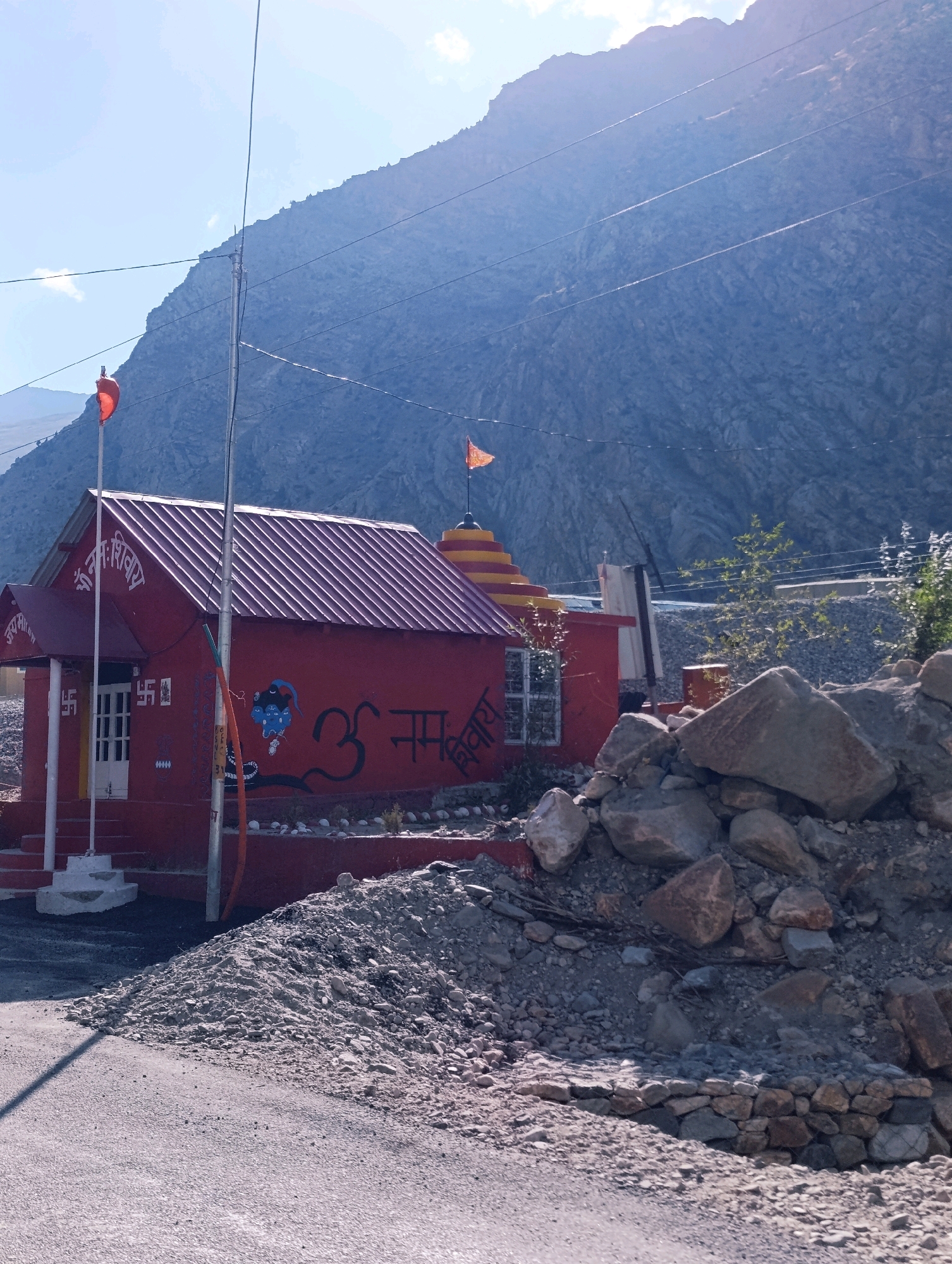 The image captures a small, red Hindu temple situated in the village of Gemur, India, nestled against a steep, rocky mountain face under a bright, hazy sky. The temple, adorned with orange and yellow tiered roofing and two flags, one orange and one red, features religious symbols such as "Om" and various other Hindi script decorations on its exterior walls. In front of the temple, there is a pile of loose rocks and debris, suggesting ongoing construction or natural erosion. A paved road runs in front of the temple, with a shadow cast across it from an unseen object. The overall scene depicts a remote, mountainous region with traditional religious architecture.