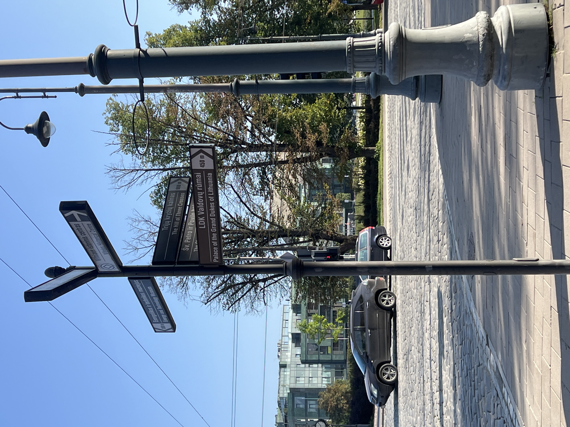The image depicts a street scene in Vilnius, Lithuania, captured on a clear, sunny day. A street signpost, a prominent feature in the foreground, displays directional information to several landmarks. Some of the visible destinations include "Vilniaus istorinis centras" (Vilnius Historical Centre), "Energetikos ir technikos muziejus" (Energy and Technology Museum), "Trijų kryžių kalnas" (The Hill of Three Crosses), "Šv. Petro ir Povilo bažnyčia" (St. Peter and St. Paul's Church), and "LDK Valdovų rūmai" (Palace of the Grand Dukes of Lithuania).
The signpost is positioned on a cobblestone street, with a modern building complex visible in the background. Two cars are present on the street: a dark-colored SUV in the left lane and a darker station wagon in the right lane. Streetlights with ornate bases are also visible along the sidewalk. The environment suggests an urban setting with well-maintained infrastructure and trees lining the street. The sky is a vibrant blue.