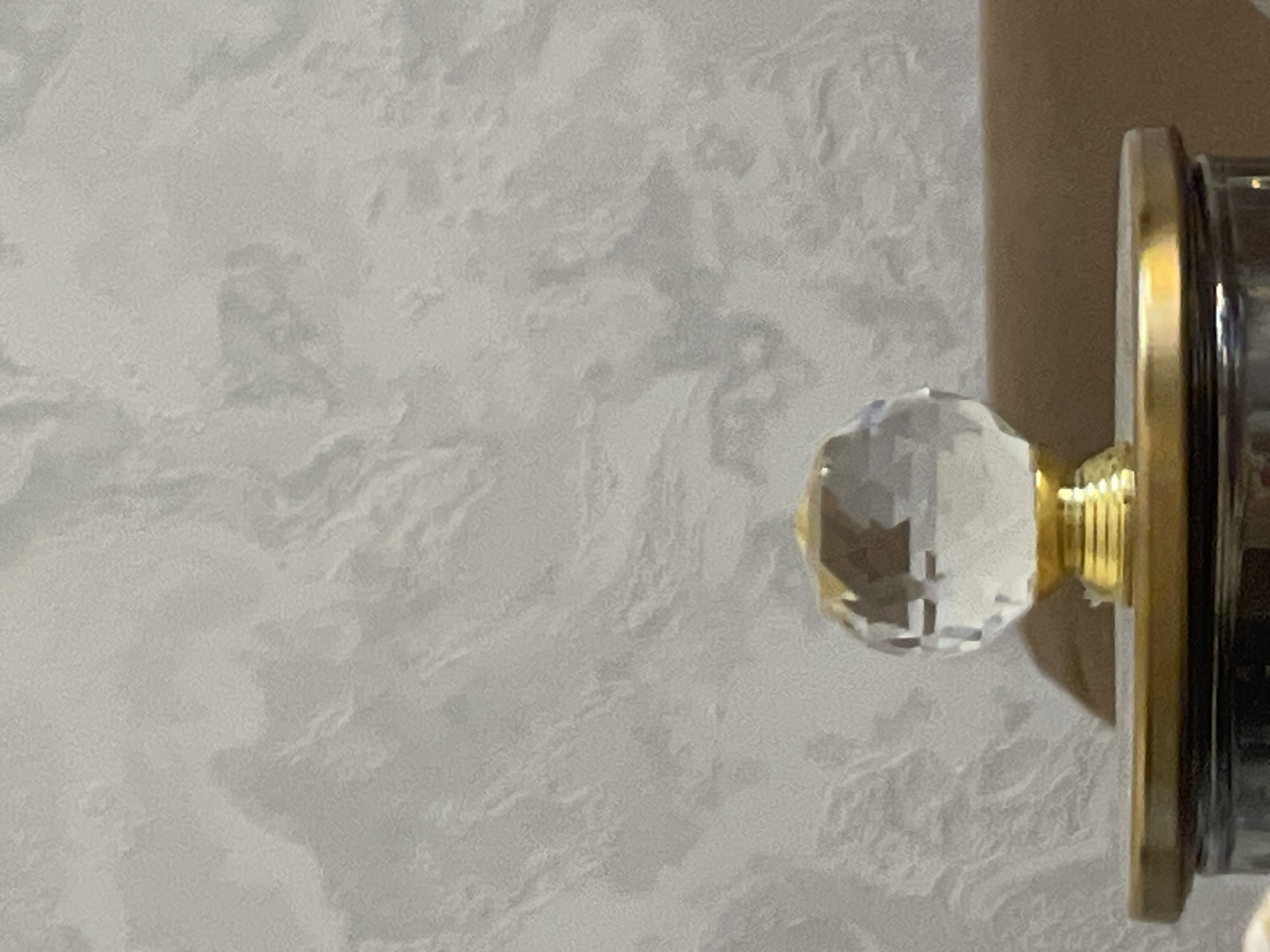A crystal knob with a golden base sits atop a dark, possibly glass, jar or container. The jar has a gold-colored rim. The knob and jar are positioned on a flat, metallic surface that is also gold-colored. In the background, a textured wall, likely wallpaper, displays a marbled pattern in shades of gray and white. The scene is indoors, with a blurred brown surface visible behind the crystal knob. The image is a close-up shot, with the focus on the knob and the immediate surface it rests upon.