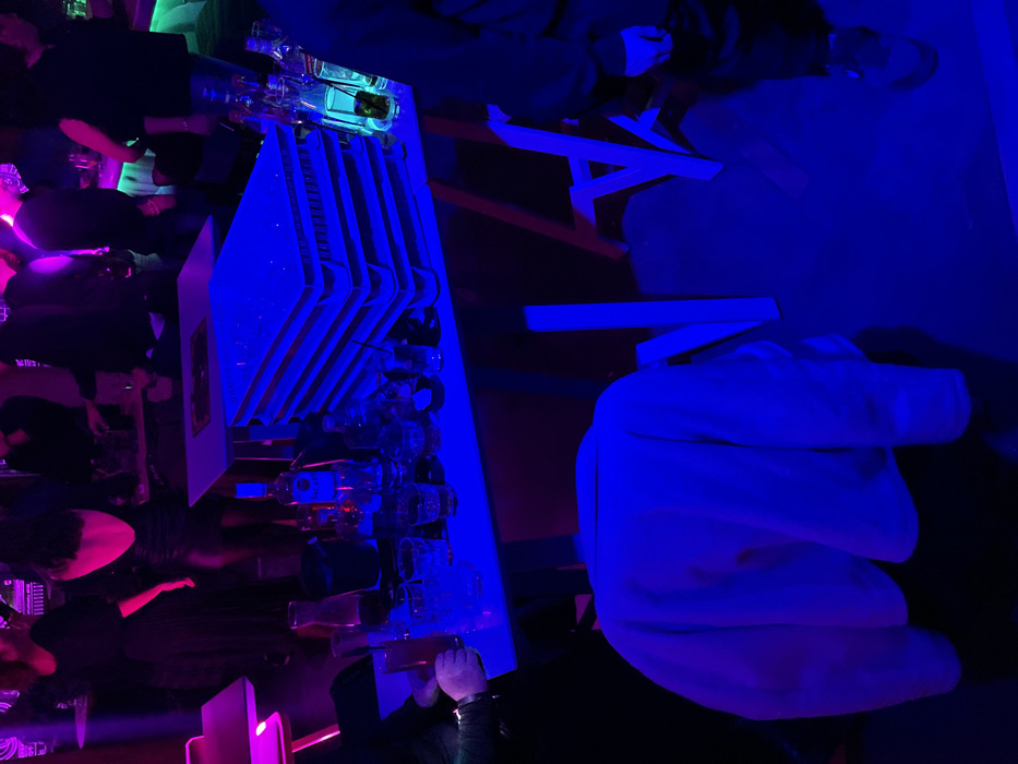 The image captures a lively scene inside a bar or club in Drapetsona, Greece, at night. The dominant feature is a long, narrow bar counter extending horizontally across the midground, brightly illuminated with intense blue neon lighting. Numerous bottles and various types of drinking glasses are neatly arranged on and behind the counter, suggesting active service. Above a section of the counter, white, rectangular panels are stacked in a decorative or functional arrangement.
Several people are visible throughout the space. In the background, a group of individuals stands and sits along the bar, their forms often appearing as dark silhouettes against the vibrant lighting. Some are illuminated by magenta or pink light sources, particularly on the left side of the frame, hinting at a dynamic lighting setup. In the foreground, partially visible on the right, a person wearing a light-colored hoodie is present, their back mostly towards the camera. Other individuals are scattered around, creating an impression of a social gathering.
The overall atmosphere is one of a bustling nightlife venue, characterized by the striking electric blue and magenta artificial lighting, which casts long shadows and creates a cool, energetic ambiance. The presence of bottles and glasses indicates an event centered around beverages. No discernible text is visible on any objects or signs within the frame.