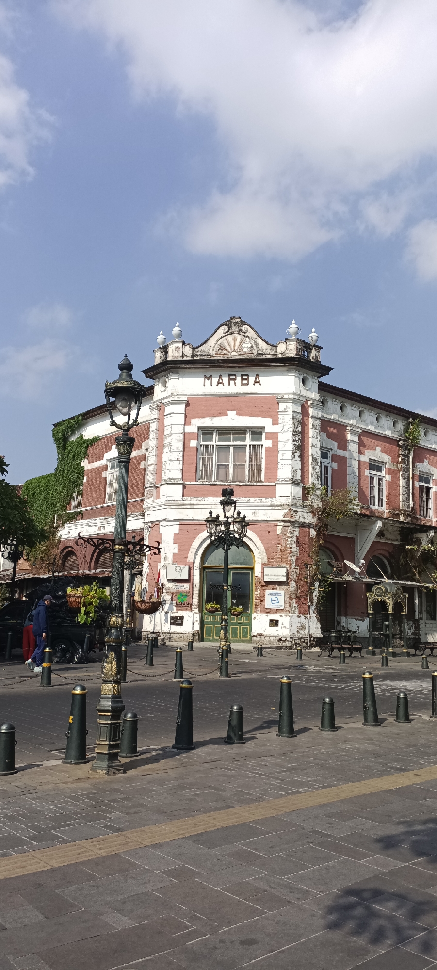 This is a vertical, eye-level, outdoor shot of the historic Marba building, located in the Kota Lama or Old Town district of Semarang, Indonesia. The building, with its distinctly colonial architecture, features red brick walls with ornate white trim and peeling paint. The word "MARBA" is clearly visible in large, dark letters across the upper facade. The building has multiple windows, some with barred lower panes and others with green shutters. A large arched doorway with green doors is visible on the right side of the building. Several streetlights with intricate metalwork stand in the foreground, casting shadows on the paved walkway and road. Black bollards with gold caps line the edge of the walkway. A person wearing a blue jacket and hat is walking on the left side of the image, and other indistinct figures and objects are visible in the background, suggesting activity in the area. Greenery, including ivy, climbs the side of the building on the left. The sky is a clear blue with scattered white clouds.