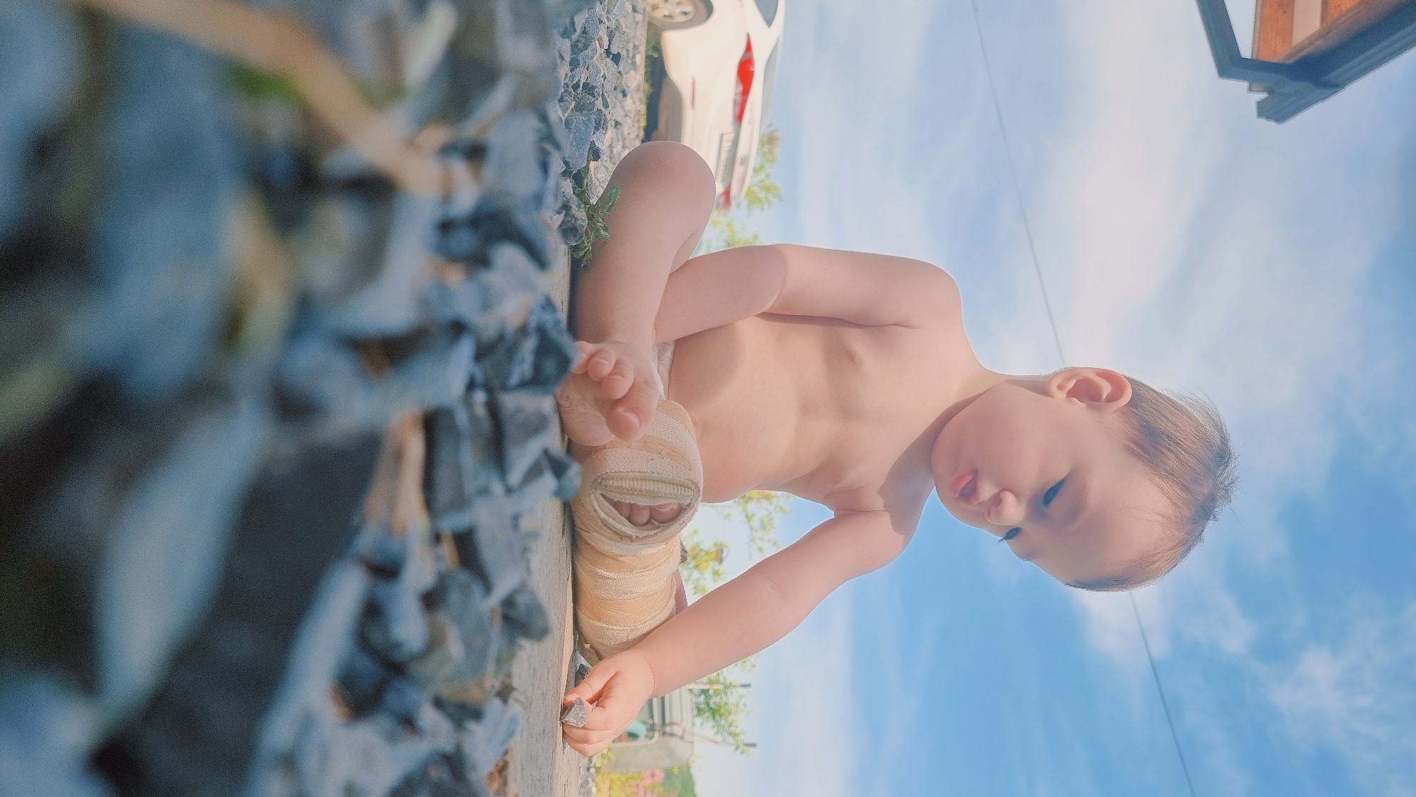 A baby sits on a gravel surface, appearing to be outside, likely in Dunnae-myeon, South Korea. The baby has a cast on one leg and is examining a small rock. Part of a white car is visible in the background. The sky is blue with some clouds. A building is also partially visible in the background.