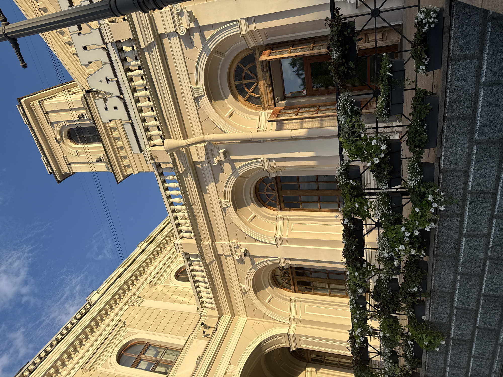 The image displays a grand architectural facade under a clear blue sky with sparse clouds. The building, likely in a classical or neoclassical style, is rendered in a warm, light cream or beige color, adorned with ornate details such as balustrades, cornices, and decorative moldings. Several windows are visible, including large arched and circular designs with elaborate wooden frames, and some rectangular ones.
Prominently featured on the upper part of the building, attached to a dark street light pole, is a large sign with the Cyrillic text "БАТ" (BAT). Strong, directional sunlight illuminates the facade, creating clear shadows that highlight the architectural depth and textures, suggesting it is daytime, possibly morning or late afternoon.
On the right side of the image, a vertical metal structure supports numerous black planters overflowing with white flowers and green foliage, forming a vibrant vertical garden. Below this, a textured gray surface, possibly a sidewalk or granite wall, shows patterns of light and shadow. Thin wires are visible stretched across the sky above the building. There are no people or specific activities taking place, contributing to a serene and majestic atmosphere. The image is oriented sideways, with the building appearing to lean into the frame from the right.