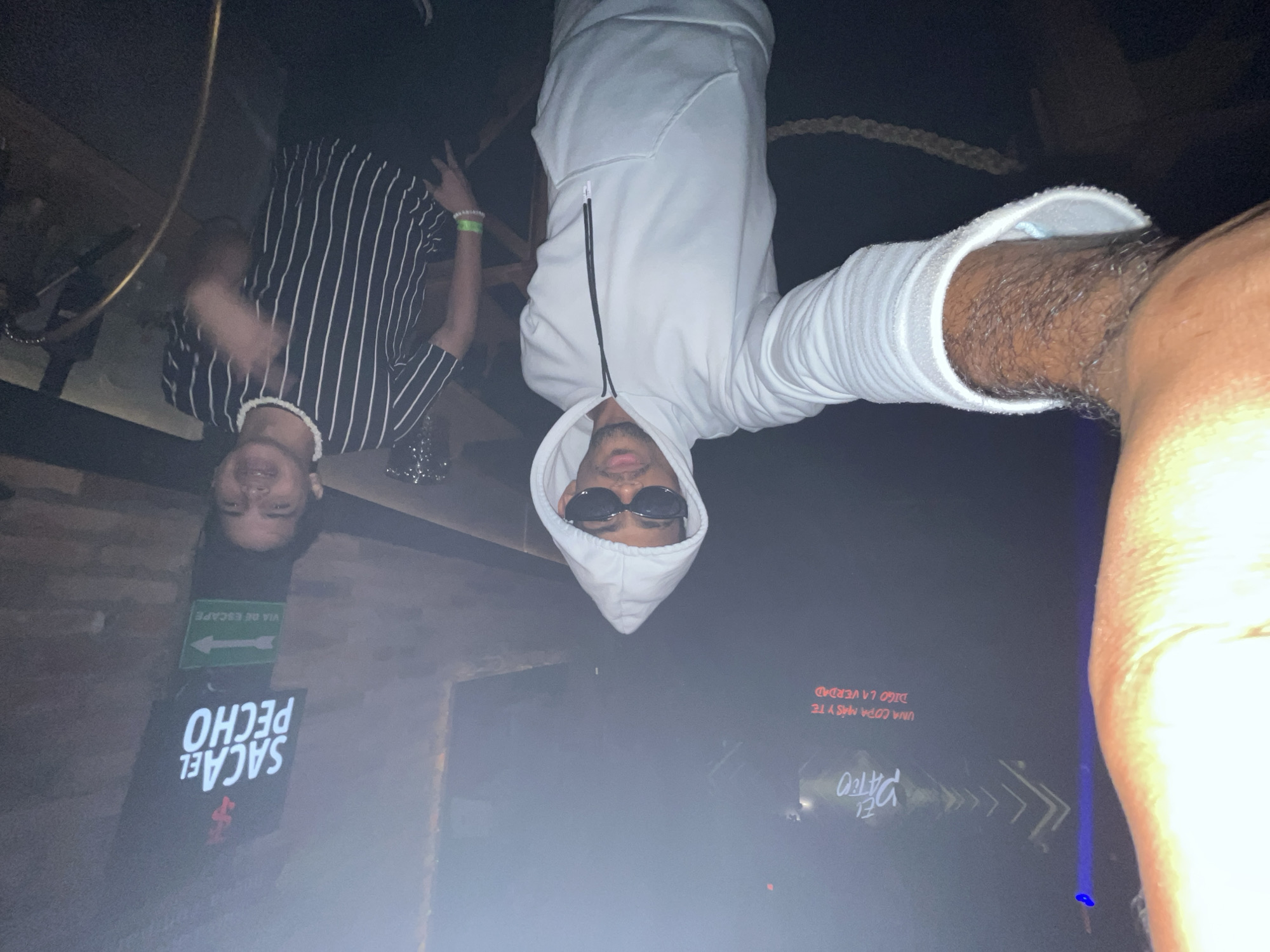 An inverted and rotated selfie captures two men inside a dimly lit, possibly hazy indoor venue, likely a club or bar in Porlamar, Venezuela, at night. The person taking the photo, positioned closest to the camera, is wearing a white hooded sweatshirt with a zipper and dark sunglasses, with a visible, hairy forearm extending towards the lens. Further back, another man is seen smiling, wearing a black and white vertical striped t-shirt and what appears to be a white beaded necklace. He also has a green wristband on his left arm.
The background reveals brickwork and various illuminated signs. One green sign, correctly oriented, reads "VIA DE ESCAPE" (Escape Route) with an arrow pointing right. Below it, a black sign displays white text, which when correctly oriented, reads "SACA EL PECHO" (Stick out your chest) next to a red logo. In the deeper background, another faint red sign, when correctly oriented, reads "UNA COPA MÁS Y TE DIGO LA VERDAD" (One more drink and I'll tell you the truth), reinforcing the bar/club setting. The overall atmosphere suggests a casual social gathering.