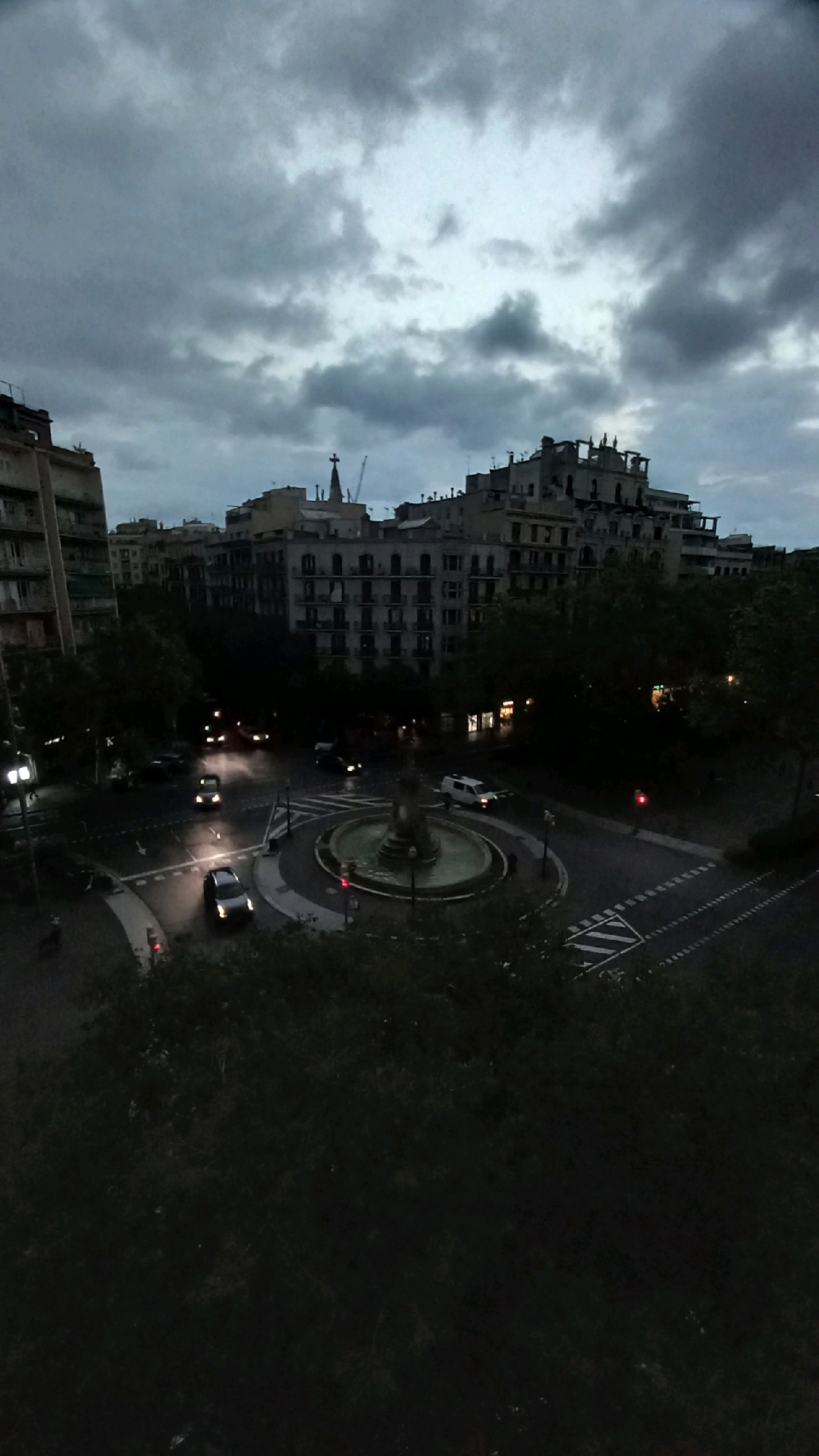 An overhead, high-angle, vertical shot captures a dusky cityscape in Barcelona, Spain, under a cloudy sky. The image focuses on a roundabout with a fountain at its center. Several cars are visible navigating the roundabout and adjacent streets, their headlights on, indicating low light conditions. The buildings surrounding the roundabout are multi-story residential and commercial structures, typical of a European city. Trees line some of the streets, their foliage appearing dark and dense in the dim light. The overall atmosphere is one of twilight or an overcast day, with muted colors and limited visibility. The scene is devoid of any visible pedestrians.