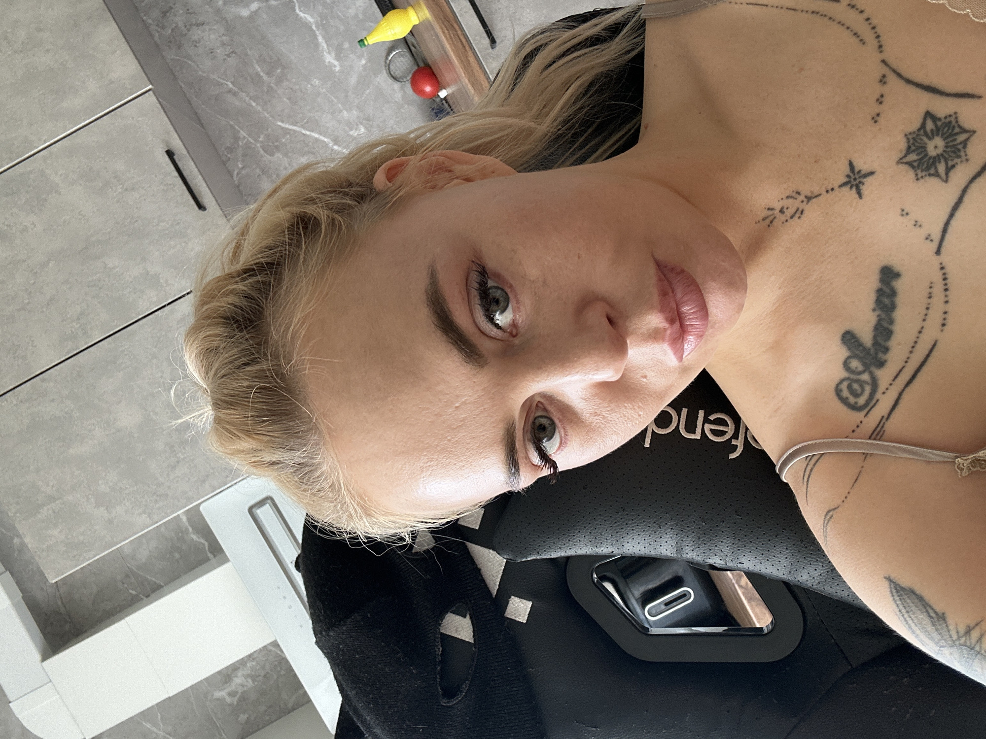 A fair-skinned adult female is depicted in a close-up selfie, looking directly at the camera with a neutral expression. Her blonde hair is styled away from her face, revealing light-colored eyes with visible makeup, including winged eyeliner and mascara. She has faint freckles or hyperpigmentation on her cheeks and wears a light-colored, thin-strapped top. Several tattoos are visible on her chest and shoulders: "Amour" in script font, a dotted line pattern, a star or mandala design, and a smaller arrow-like motif on her sternum, along with a partial feather or leaf design on her left arm.
She is seated in what appears to be a black gaming chair, identifiable by its textured surface and a partial "Defender" logo. The background reveals a modern kitchen or utility space. To the upper left, a white wall-mounted range hood or cabinet is visible against a grey tiled wall with a subtle marble-like pattern. Above and to the right, grey kitchen cabinets with black horizontal handles are mounted. A wooden countertop is visible on the right, displaying a red spherical object, a metallic ring, and a yellow lemon-shaped squeeze bottle. The overall environment suggests a contemporary indoor setting in the city of Unknown, Unknown.