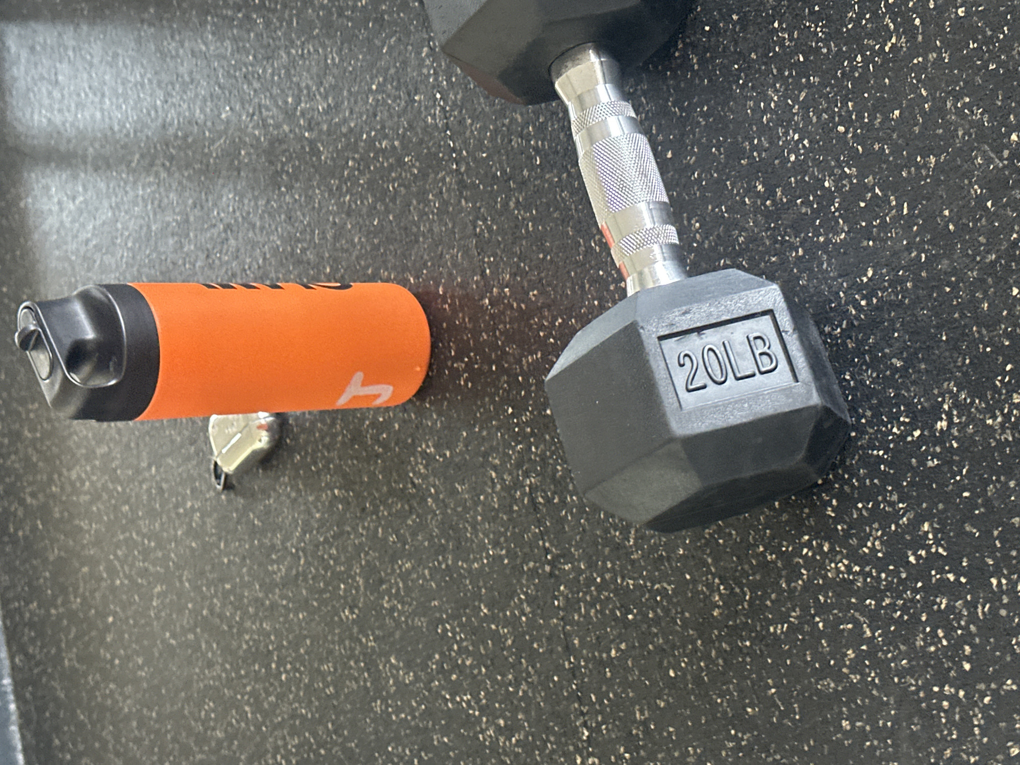 The image shows a 20-lb dumbbell and an orange water bottle on a dark gray speckled floor, likely in a gym setting. No people are visible. The exact location in Huixquilucan de Degollado, Mexico, cannot be determined from the image.
