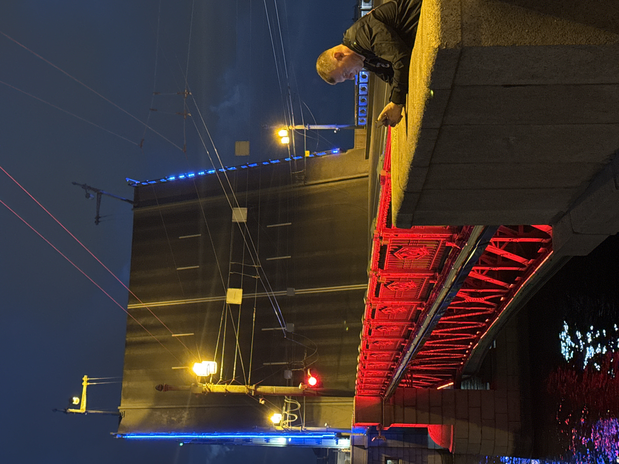 An individual in a dark jacket is seated on a stone ledge, looking down at a smartphone. The scene takes place at night in Saint Petersburg, Russia, near a bridge that is raised and illuminated with blue and yellow lights. A red-lit pedestrian walkway is visible in the foreground, spanning over dark water reflecting the lights. The sky is dark and cloudy.