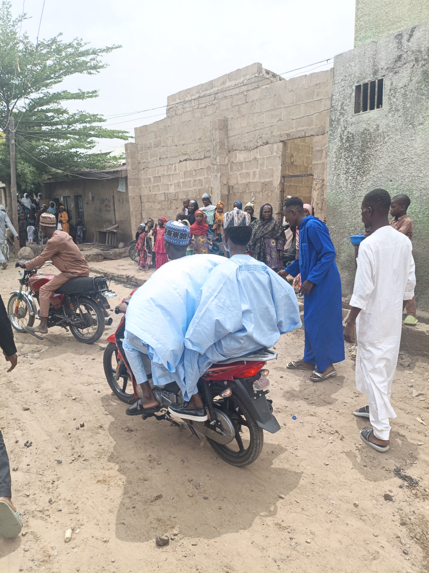 The image depicts a street scene in Gwagwarwa, Nigeria. In the foreground, two men are on a red motorcycle. The driver, wearing a light blue garment and a patterned cap, is facing away from the camera, bending forward. The passenger, also in a light blue garment, is seated behind the driver. In the mid-ground, a man in a tan outfit is riding a red motorcycle, looking to the right. A crowd of people, including women and children in colorful clothing, are gathered in the background, some observing the motorcyclists. The environment appears to be a dusty, unpaved street lined with buildings constructed from concrete blocks. A large tree is visible on the left. The sky is overcast.
