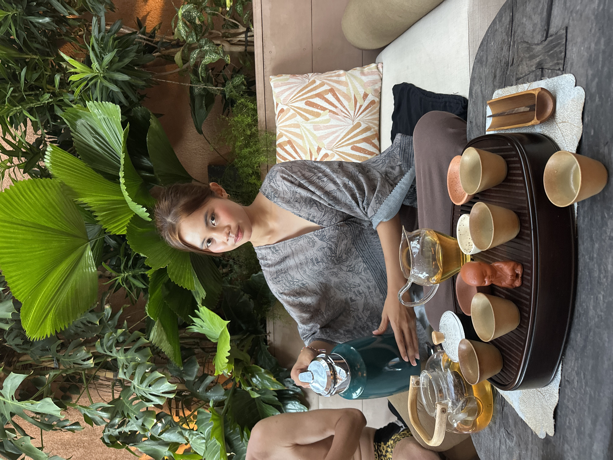 The scene depicts a woman in Pecatu, Indonesia, seated on a couch surrounded by lush greenery. She is positioned near a low wooden table where she is conducting a tea ceremony. The table holds a tray with several small teacups, teapots, and other tea-related objects. The woman is pouring liquid from a dark green thermos into one of the teapots. The environment is natural and tropical, with various plants and a patterned pillow adding to the relaxed atmosphere. There is also another person, possibly a man based on the partial view of his bare chest and patterned clothing, sitting beside her.