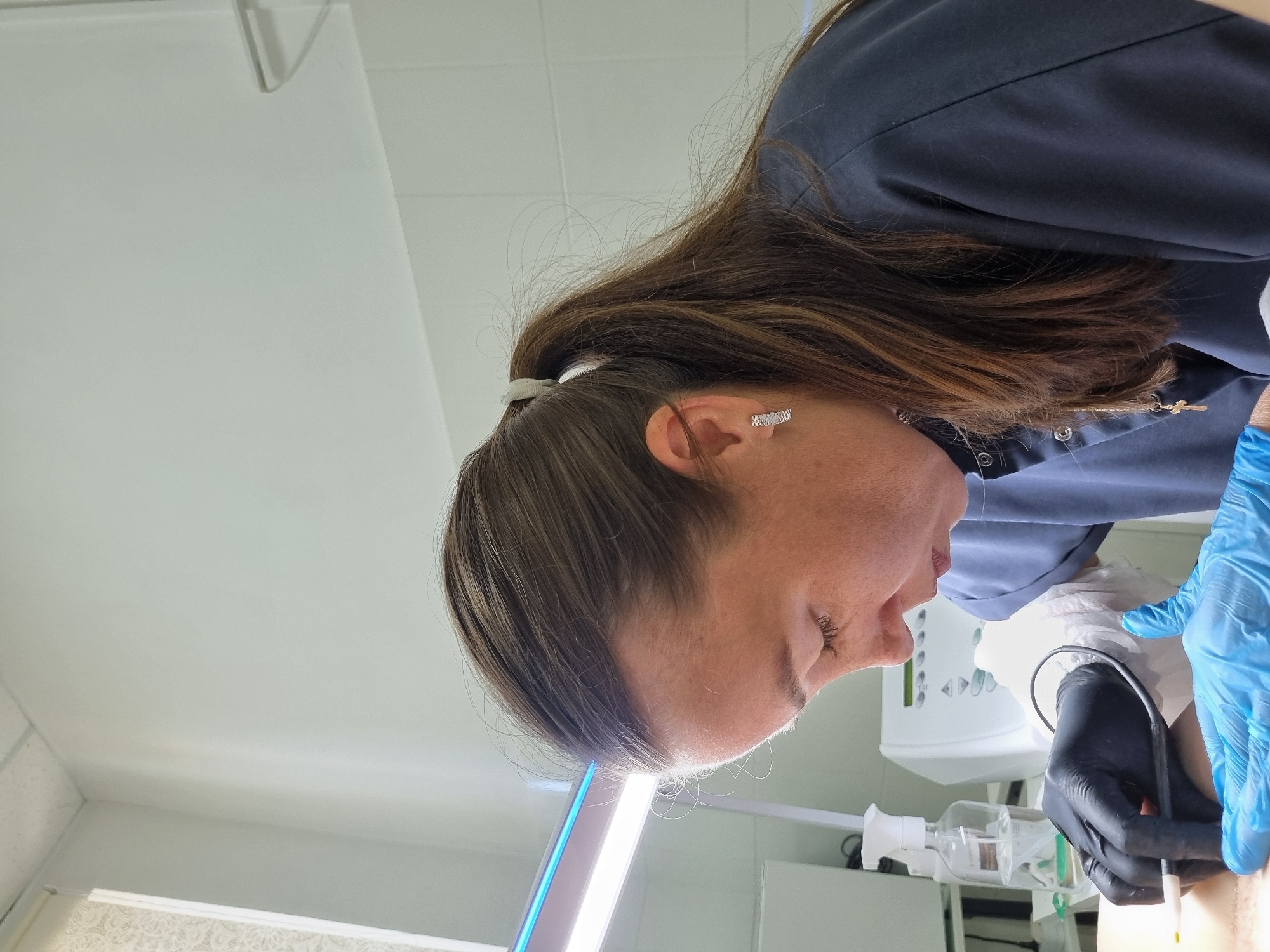 The image shows a close-up, side profile of a woman with brown hair pulled back in a ponytail, wearing a dark blue scrub top. She is wearing black gloves on her left hand and blue gloves on her right hand. Her left hand is holding a device with a circular cord, and her right hand is holding another instrument near what appears to be a person's skin. She is looking down intently at her work. In the background, there is a bright blue light fixture, a white machine, and a spray bottle on a shelf. The environment appears to be a medical or cosmetic treatment room. The image does not contain any information to determine the location in the city of Chaikovsky, Russia.