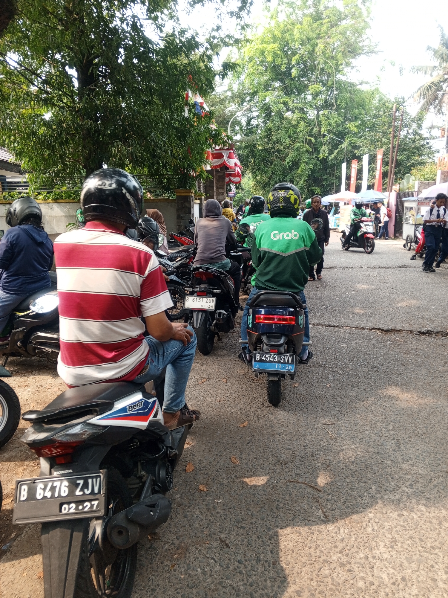 In Depok, Indonesia, several individuals are gathered on a street, primarily on motorcycles. The scene appears to be an outdoor marketplace or a gathering point, with trees and some temporary structures visible in the background. A few people are wearing the distinctive green jackets of Grab, a ride-hailing service. One individual in the foreground is seated on a motorcycle, wearing a red and white striped shirt and blue jeans, with a black helmet. Another person in a green Grab jacket is also on a motorcycle in the mid-ground. Further back, a person wearing a gray hoodie is on a motorcycle, and other individuals are seen walking and standing. License plates on the motorcycles are visible, with one reading "B 6476 ZJV 02.27". The road surface is asphalt with some cracks. The lighting suggests it is daytime, with sunlight casting shadows.