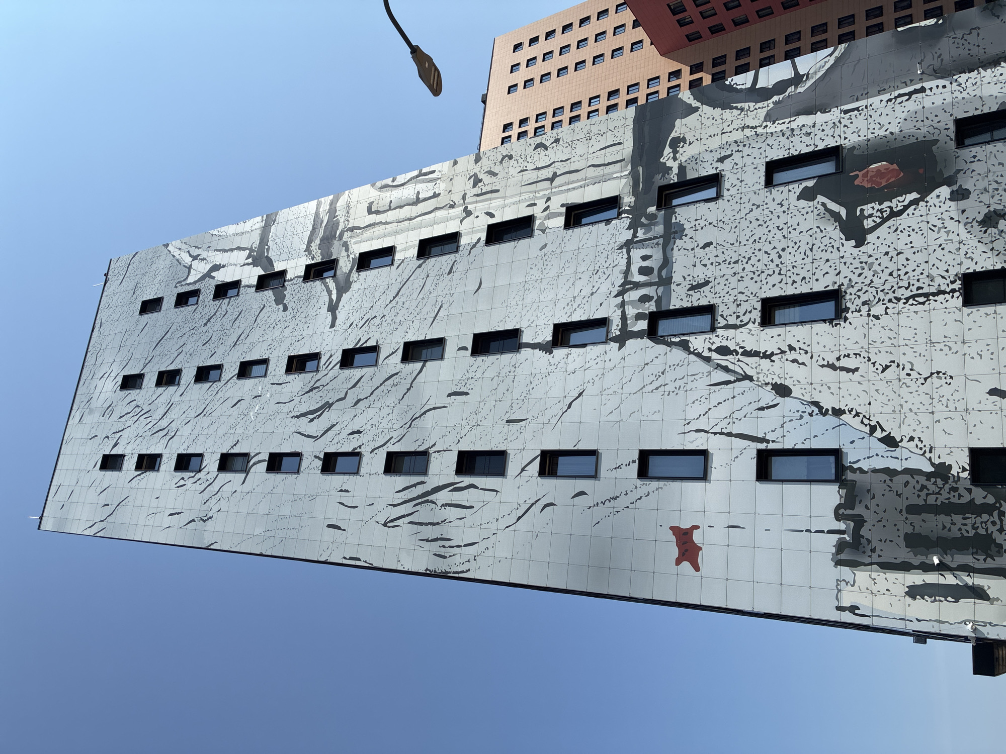 This is a low-angle, eye-level exterior shot of a modern, light-gray building facade decorated with a large black and white mural. The mural features abstract, organic shapes, and what appears to be a stylized depiction of a castle or fortress with a red flag. The building is tall and narrow, with numerous dark-framed rectangular windows evenly spaced across its surface. The sky is clear and blue. In the background, to the right, are parts of two other modern buildings: one is a light-orange building with many windows, and another is a darker red building. A street lamp with a curved arm extends from the upper right of the frame, casting a shadow on the mural. The building with the mural is located in Kommunarka, Russia. There are no people visible in the image.