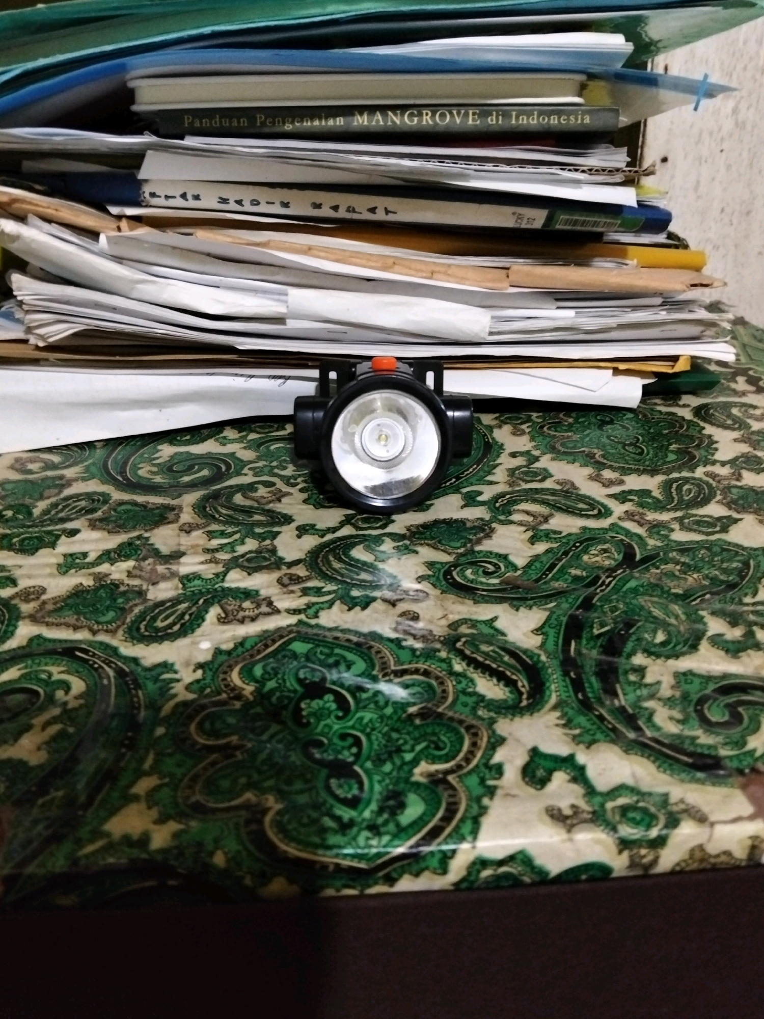 A headlamp is placed on a table covered with a green and beige paisley patterned cloth. Behind the headlamp, a stack of papers and books is visible. The items are arranged on a flat surface. The image appears to be taken indoors. The overall scene suggests a workspace or a personal study area. The presence of books and papers could indicate an educational or research setting. The paisley pattern on the table adds a decorative element to the otherwise utilitarian arrangement of objects. The lighting suggests an indoor environment, possibly with artificial light.