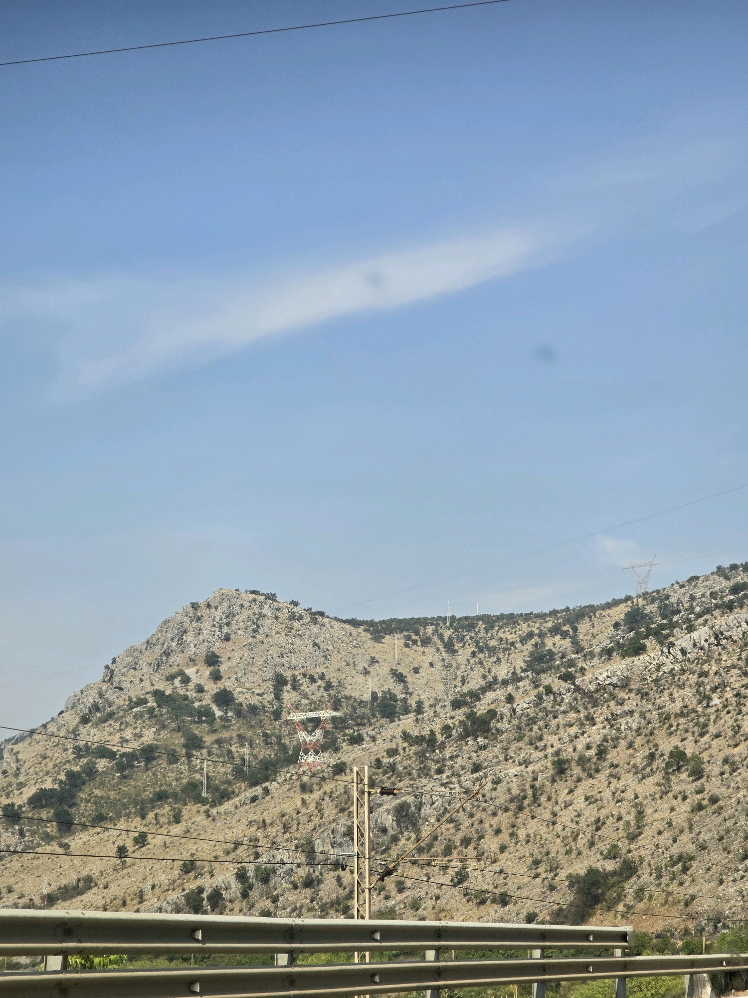 The image shows a mountainous landscape under a clear blue sky with a few wispy clouds. In the foreground, a metal guardrail runs horizontally across the bottom of the frame. Behind the guardrail, the slope of a dry, rocky mountain is visible, dotted with sparse green vegetation. Several power lines and pylons are present on the mountain and in the sky, with one distinctive red and white lattice pylon prominently featured. The environment suggests a rural or semi-rural area with infrastructure for electricity transmission. The scene is likely taken from a moving vehicle on a road, given the perspective and the presence of the guardrail. Without specific landmarks, pinpointing an exact location within Podgorica, Montenegro is not possible from the image alone, but the mountainous terrain is characteristic of the region surrounding the city.