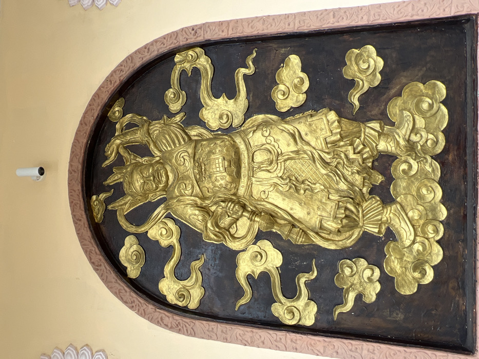 The image displays a large, golden bas-relief sculpture embedded within an arched recess on an exterior wall of a building in Đăk Mar, Vietnam. The central subject is an elaborately armored deity with a stern expression, wearing a helmet or crown, and holding a staff-like weapon in its raised right hand. The figure stands on golden clouds amidst a dark brown background adorned with additional golden cloud motifs. The arched frame surrounding the sculpture is reddish-brown with carved ornamentation. The wall immediately surrounding this recess is a uniform light beige. A white cylindrical security camera is mounted on the light beige wall directly above the arch's apex. A white decorative architectural element is partially visible on the left side of the scene.