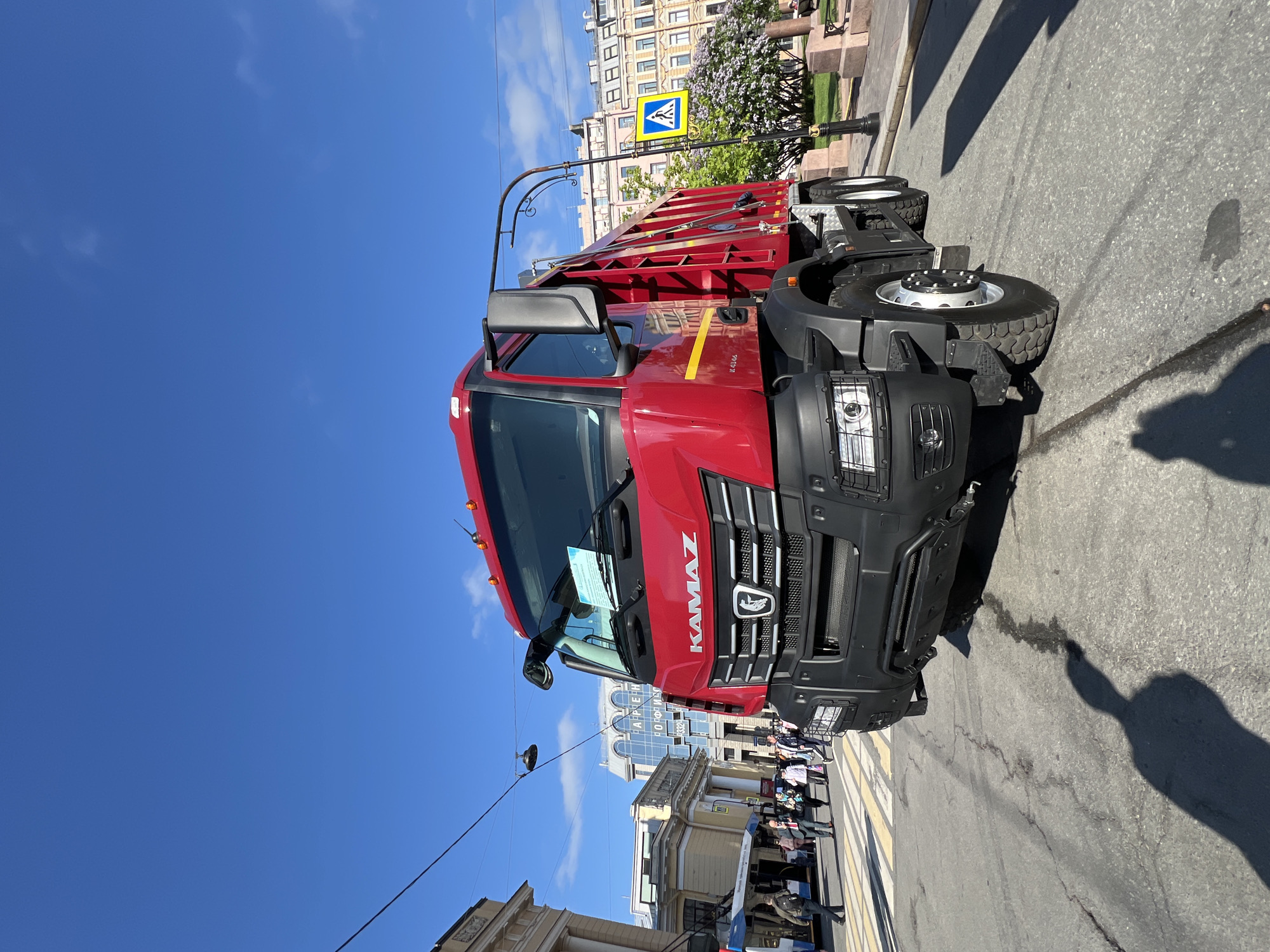 A red KAMAZ truck is parked on a city street in what appears to be Saint Petersburg, Russia. The truck is positioned in front of a building, partially obscuring its view. A crowd of people can be seen walking on a zebra crossing nearby. A pedestrian crossing sign is visible near the truck. The sky is clear and blue. The shadow of the person taking the photo is cast on the street.