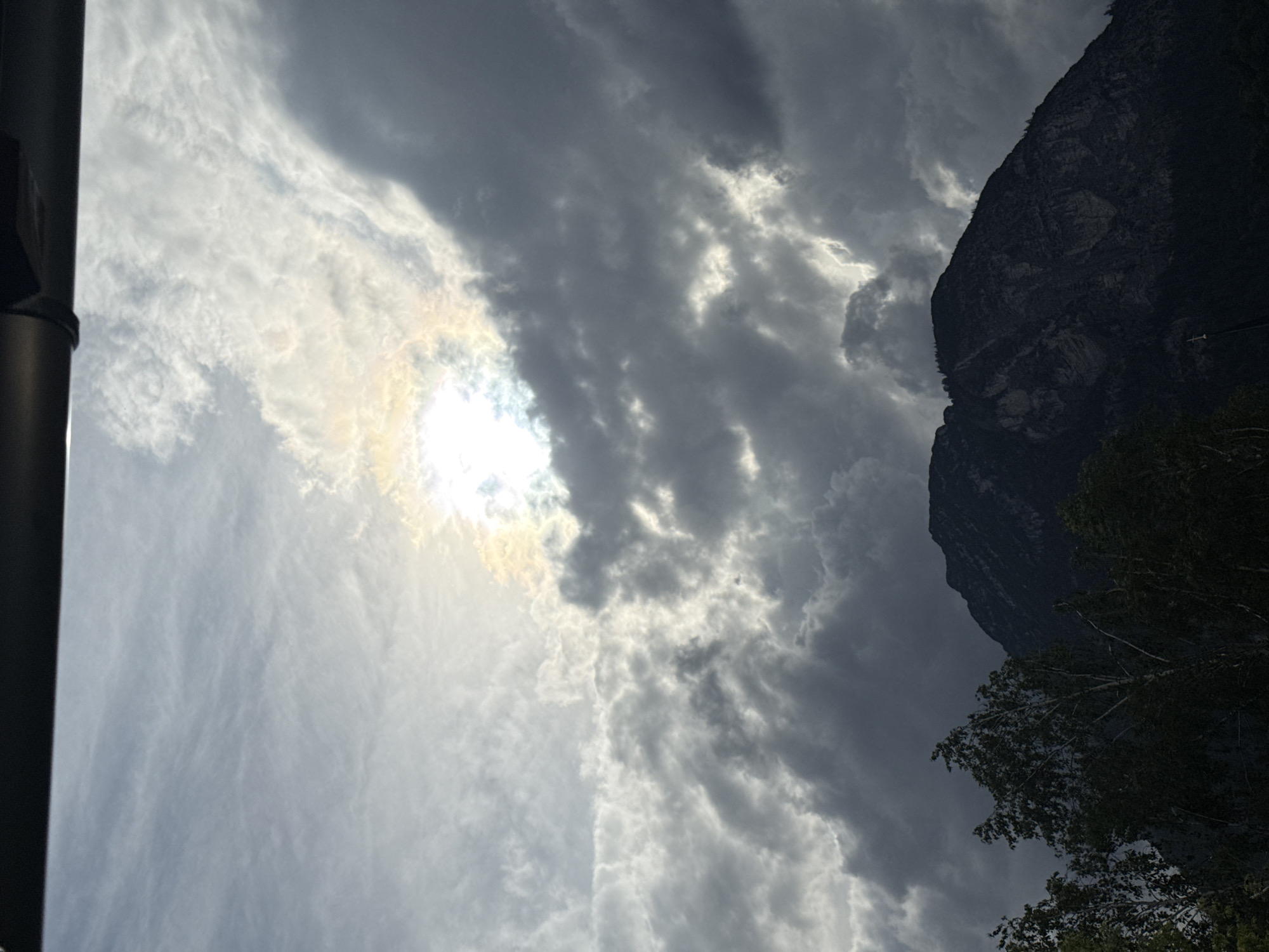 The image shows a dramatic sky above a dark mountain range, with the sun peeking through a break in the clouds, creating a halo effect. The mountain appears to be the Wank mountain in Garmisch-Partenkirchen, Germany. The foreground is filled with dark, silhouetted trees. The overall impression is one of a moody, atmospheric landscape.