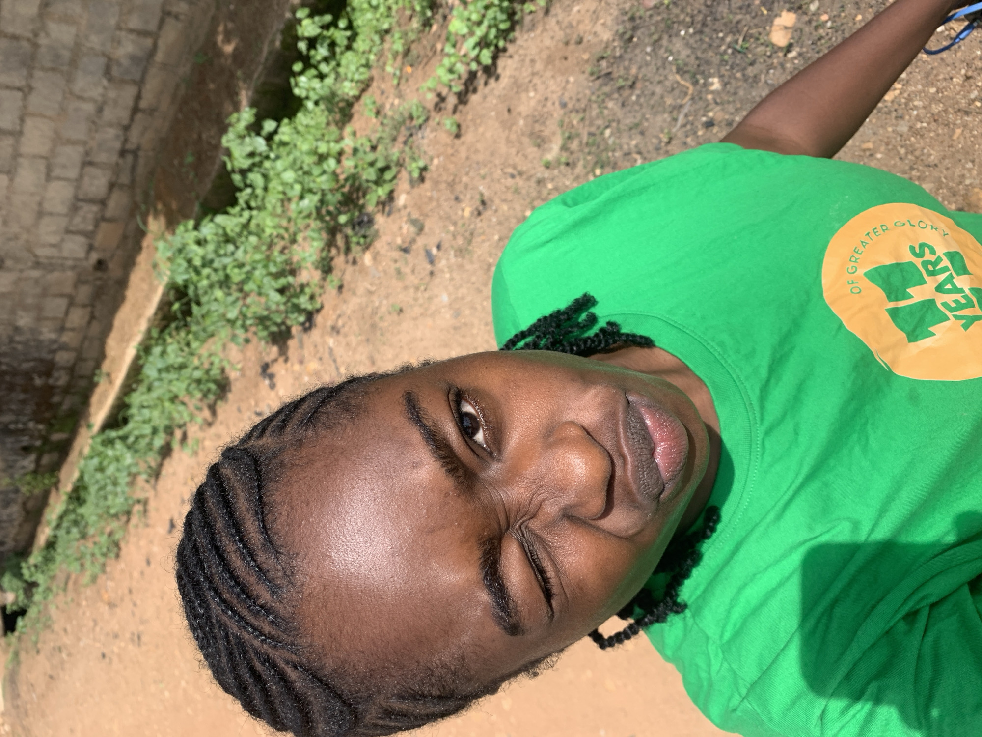 A close-up, high-angle portrait shot features a person with dark skin and braided hair, looking up towards the camera. The subject is wearing a bright green t-shirt and has an expressive face, with one eye appearing to squint or wink. Their dark braided hair is visible, pulled back from the face. The lips are slightly parted.
In the background, out of focus, is an outdoor setting. To the left, a textured stone or brick wall runs diagonally. Below the wall and extending across the middle ground, there's a patch of green foliage or small plants growing on what appears to be dry, reddish-brown earth or dirt. The lighting is bright and sunny, indicating daytime.
On the front of the green t-shirt, a circular orange logo is visible. Partially legible text on the logo reads "OF GREATER GLORY" and "YEARS," with what looks like a large "11" in the center.
The scene suggests a casual outdoor moment, possibly in Rafin Par, Nigeria, given the provided context. The overall mood is bright and informal.