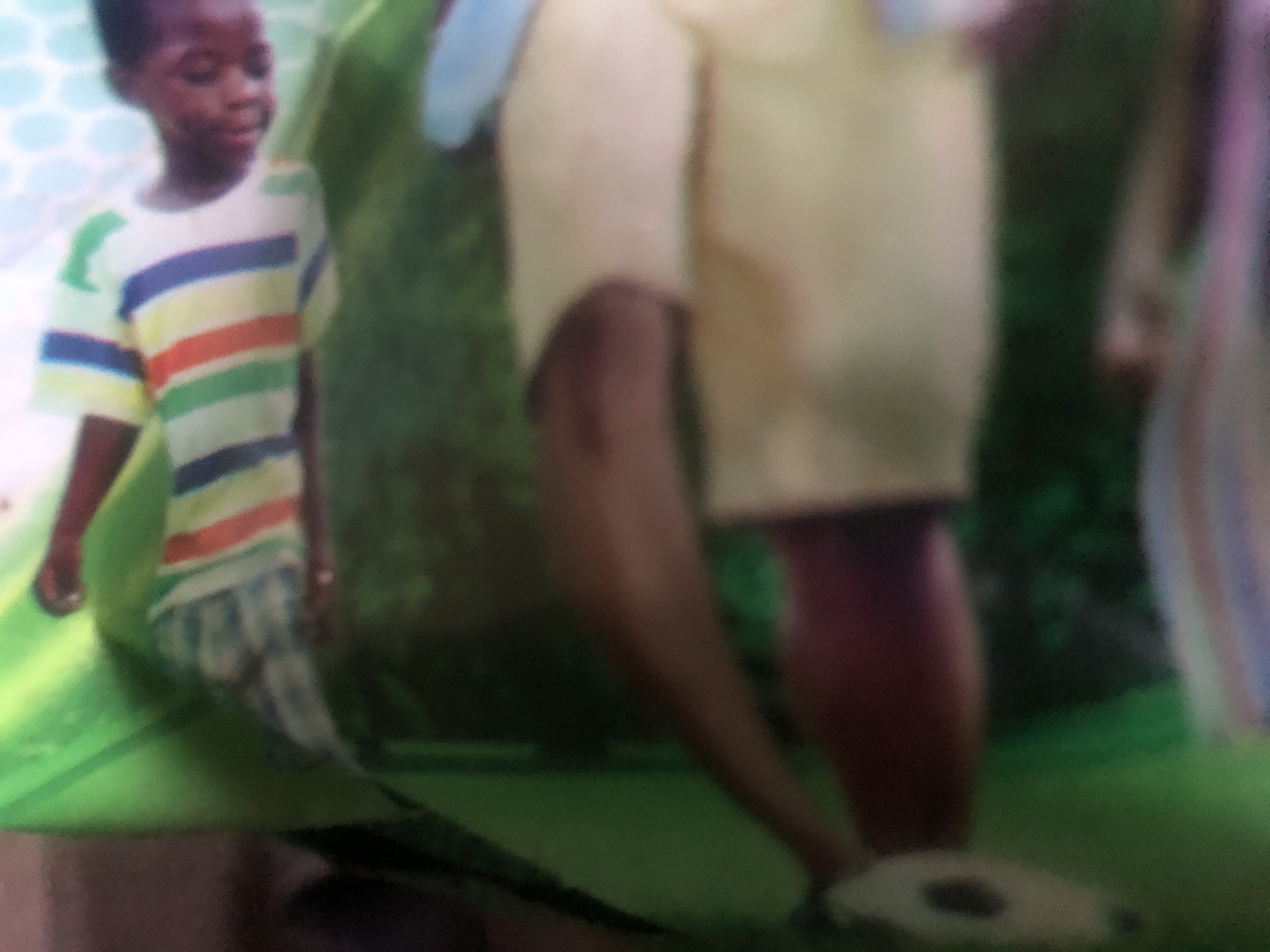 The image shows a close-up of a child wearing a striped shirt, lying down. The background is blurry and appears to be a green, grassy surface, possibly a lawn or park. There are some out-of-focus circular shapes in the background, which might be part of a toy or decoration. The overall impression is that of a playful or relaxed moment captured in a casual setting. No specific location in Jalingo, Nigeria, can be identified from the visual information provided.