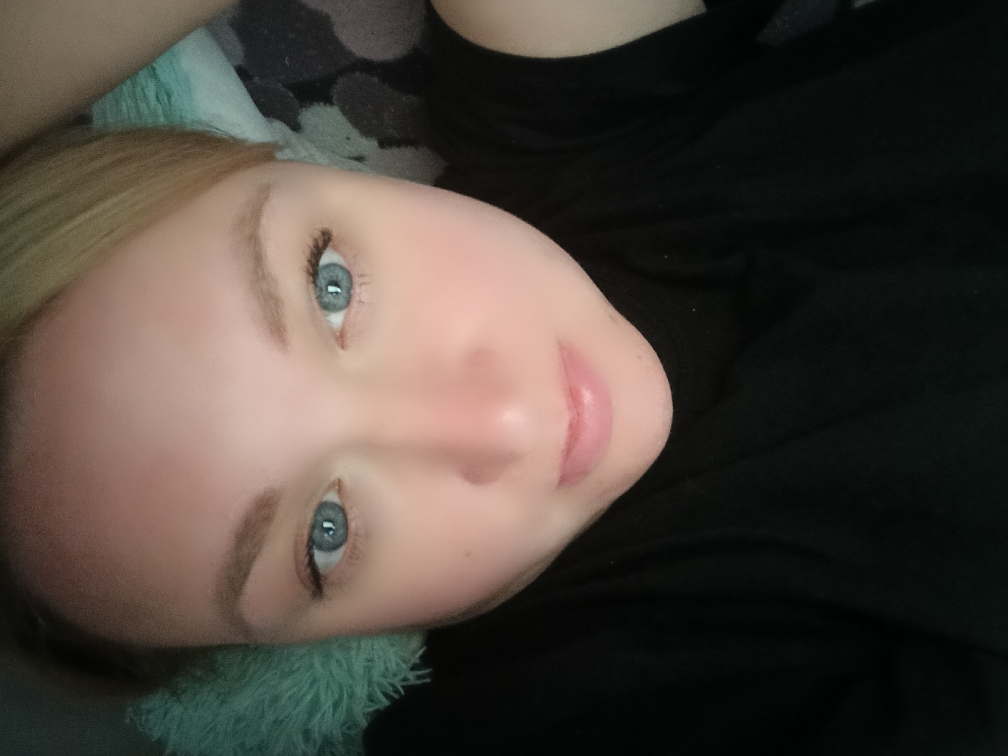 The image shows a light-skinned person with blue eyes and blonde hair lying down. The person is wearing a black shirt. A soft, light green and gray patterned surface is behind their head. There is no information available to determine the location as Chaikovsky, Russia, or any other specific location.