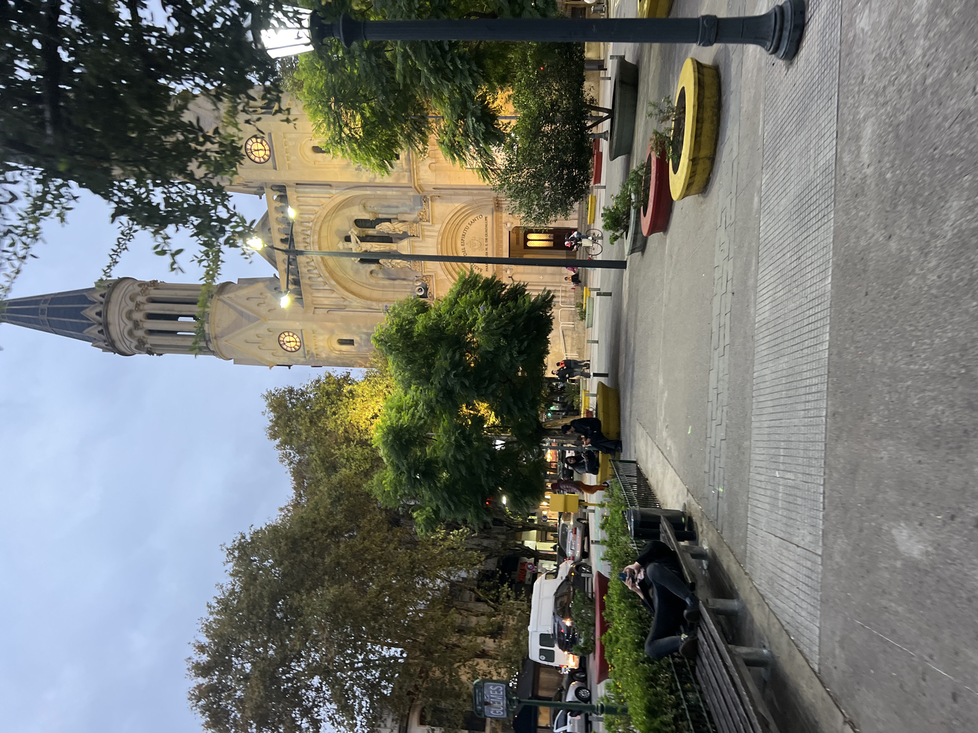 The image shows the Basilica of the Holy Sacrament in Buenos Aires, Argentina, at dusk. The basilica is illuminated, with its tower and facade prominently visible. In front of the basilica is a sidewalk with street lights, trees, and benches. People are sitting on the benches and walking along the sidewalk. Cars and vans are parked on the street alongside the sidewalk. Flower pots in red and yellow rings add color to the landscape. The sky is overcast. The overall atmosphere is calm and urban.