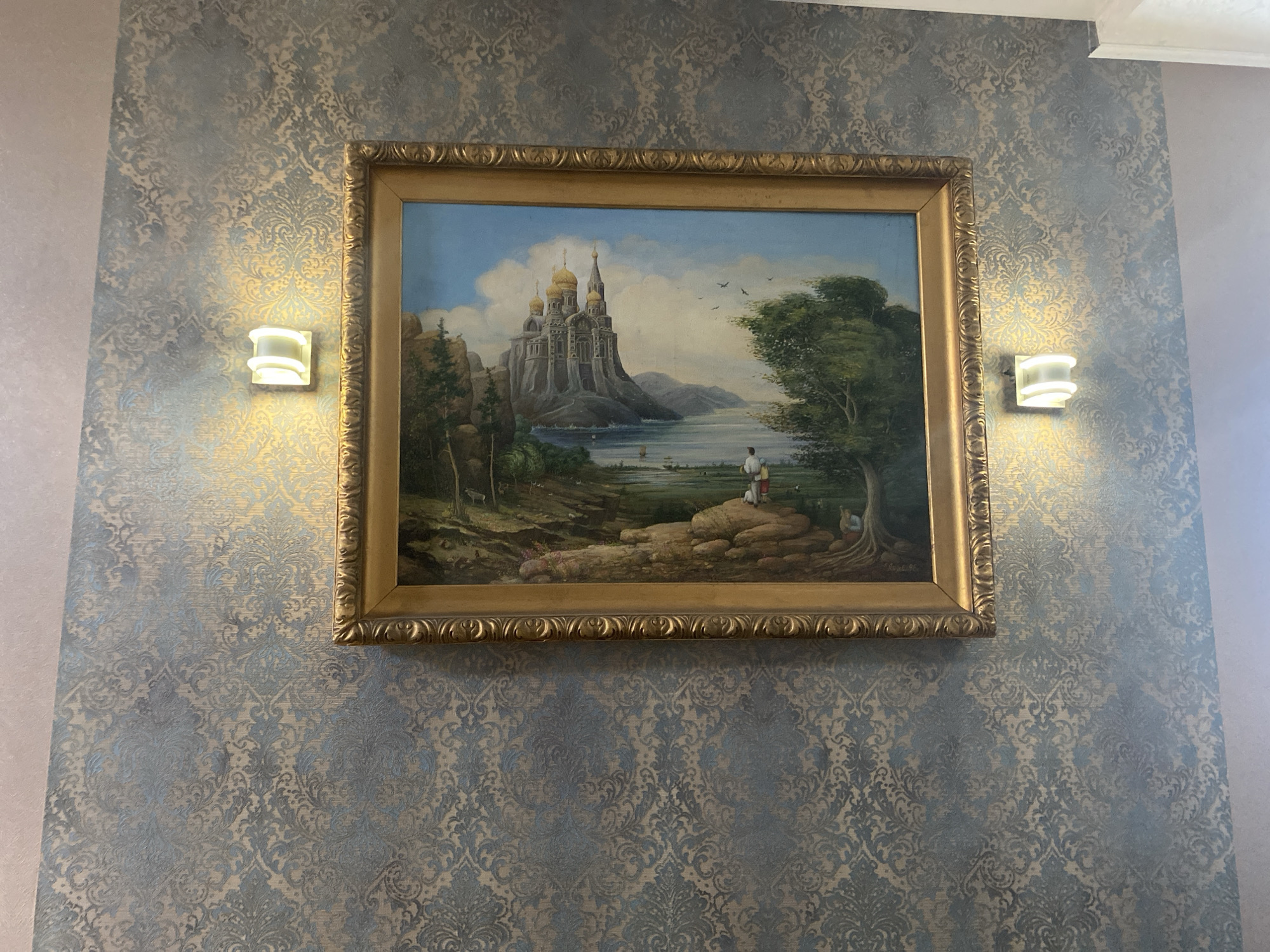 This media file features a framed painting hanging on a wall covered in ornate, damask-patterned wallpaper. The painting depicts a dramatic landscape with a large, intricate castle-like structure perched on a rocky outcrop overlooking a body of water. Golden-domed towers rise from the castle, suggesting a religious or historical building. In the foreground, two figures stand on a rocky shore, one appearing to be a man in traditional attire, and the other a smaller figure, possibly a child. Lush green trees frame the scene on the right, and smaller trees and rocks dot the landscape. The sky is a bright blue with scattered white clouds, and a few birds are visible. The overall impression is of a serene and somewhat majestic scene, possibly set during daylight. Two modern, wall-mounted light fixtures with curved shades flank the painting, casting a warm glow on the wall and illuminating the artwork. The wallpaper itself is a blend of light blue and gold, creating a textured and elegant background.
