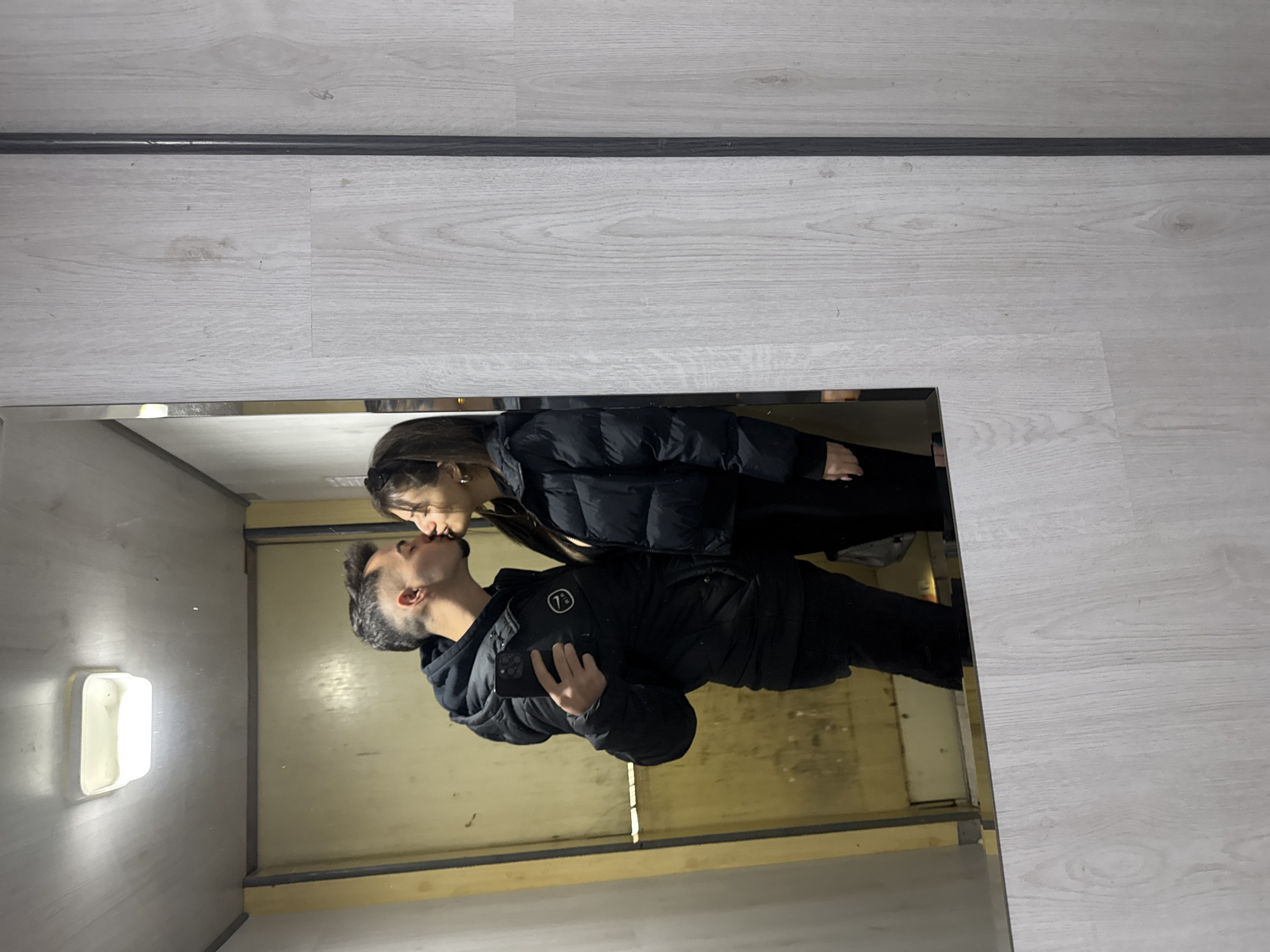 A photo shows a mirror reflection of a kissing couple, implying that the photo itself is taken from the outside of a mirror. The location is a small elevator with light cream walls and a single, rectangular light fixture. The female is on the right, with dark hair and a black puffy jacket. The male is on the left, with a dark beard, dark hair, and a black puffy jacket. Both are touching lips. The actual setting is likely on a white wooden floor based on what is surrounding the mirror, not the city of Sofia.