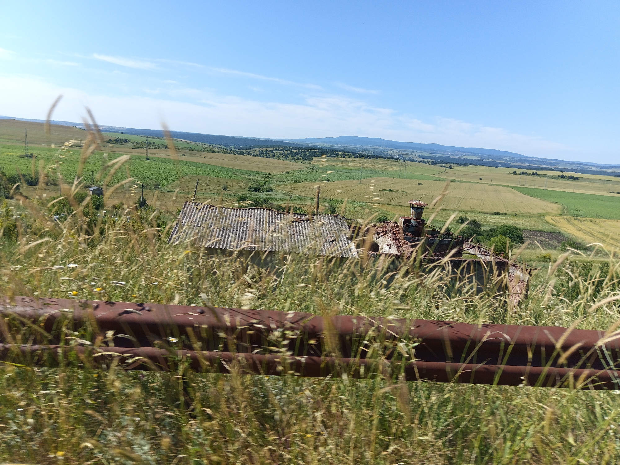The image depicts a rural landscape in Malak Manastir, Bulgaria, characterized by rolling green hills and fields under a clear blue sky with wispy clouds. In the foreground, tall, dry grasses sway, partially obscuring a rusted metal barrier. Behind the grass and barrier, the remnants of an old building with a corrugated metal roof and crumbling brick walls are visible. The building appears derelict and abandoned. Further in the distance, the landscape unfolds with more agricultural fields of varying shades of green and yellow, interspersed with trees and rolling hills that extend towards the horizon. Electrical poles are scattered throughout the fields. The overall impression is one of a quiet, somewhat desolate, rural setting.