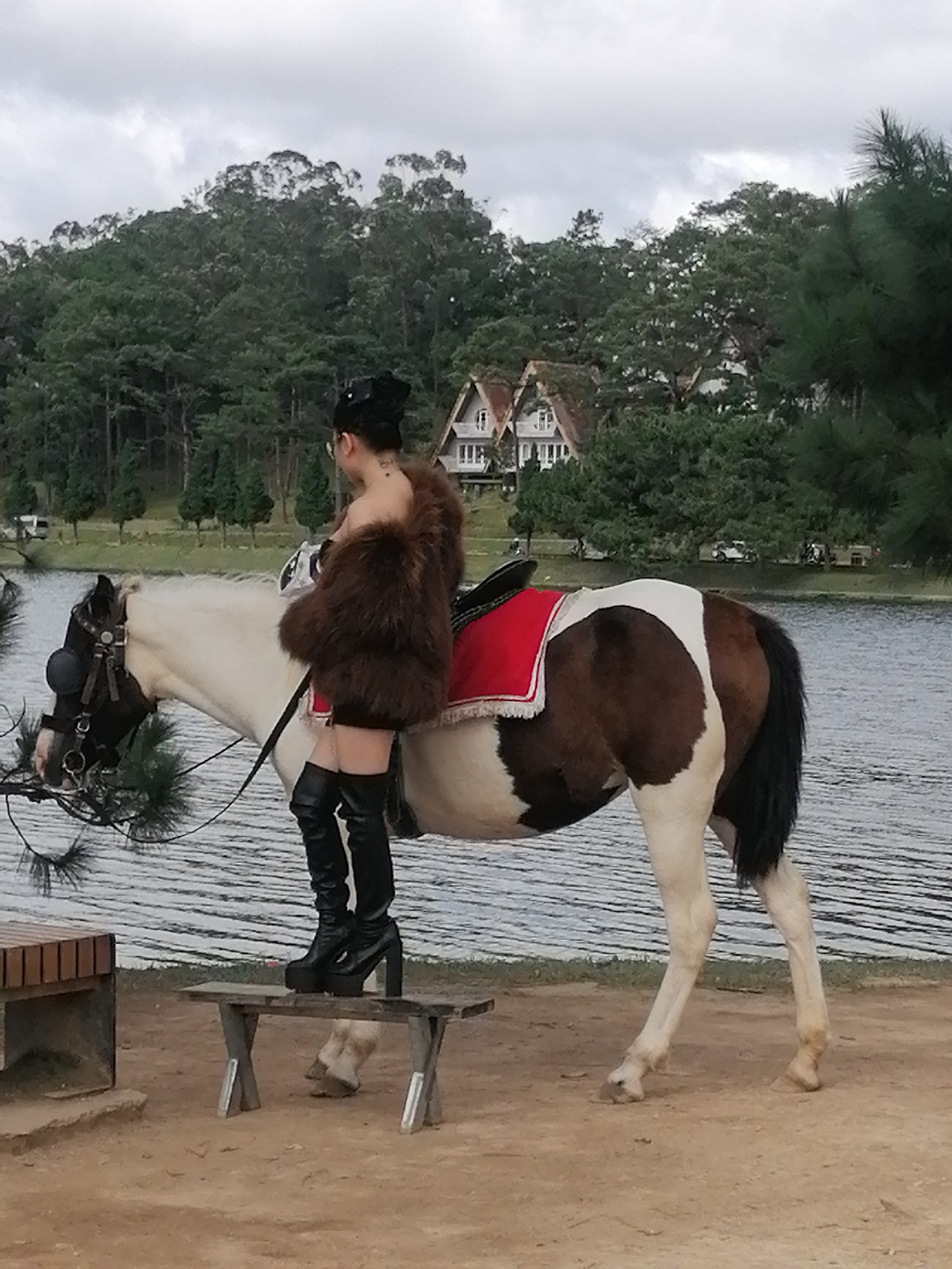A woman wearing a black hat, a brown fur coat, and black thigh-high boots stands on a small wooden bench next to a pinto horse. The horse is white with brown patches and is wearing a bridle and a red saddle pad. The scene appears to be outdoors, with a body of water in the background, likely Xuan Huong Lake in Da Lat, Vietnam, given the architecture visible in the distance. The background also features a dense forest and a cloudy sky.