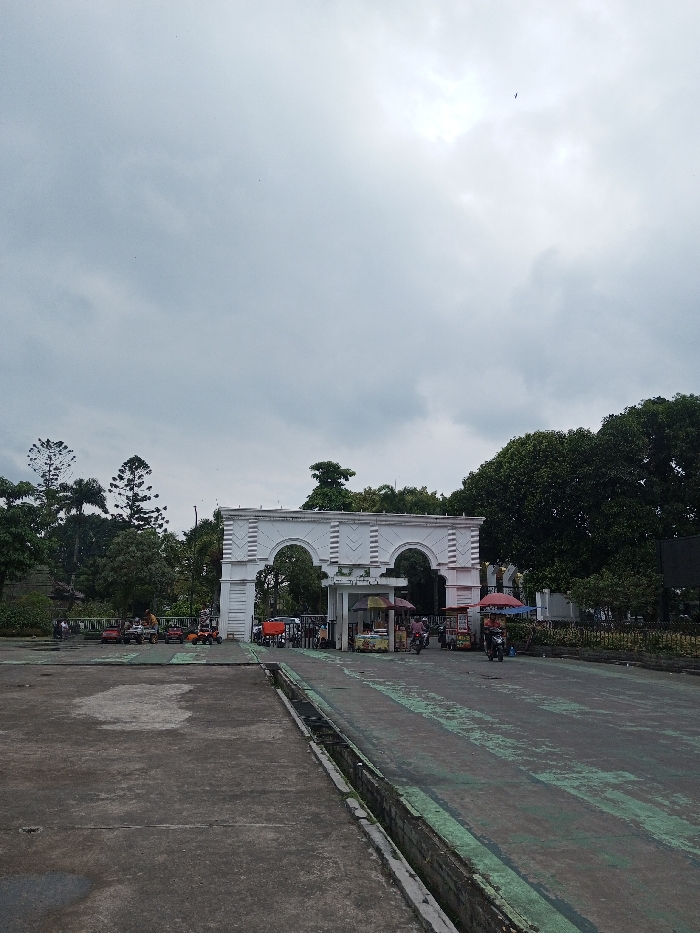 A white, ornate arched gate dominates the scene, with two symmetrical arches and supporting pillars. Trees surround the gate, some framing the structure, while others form a dense backdrop. The sky is overcast and gray. In front of the gate is a paved area with a long drain running along the left edge. There are stalls selling food to the right of the gate and there are people standing at the stall. Small vehicles are visible on the left.