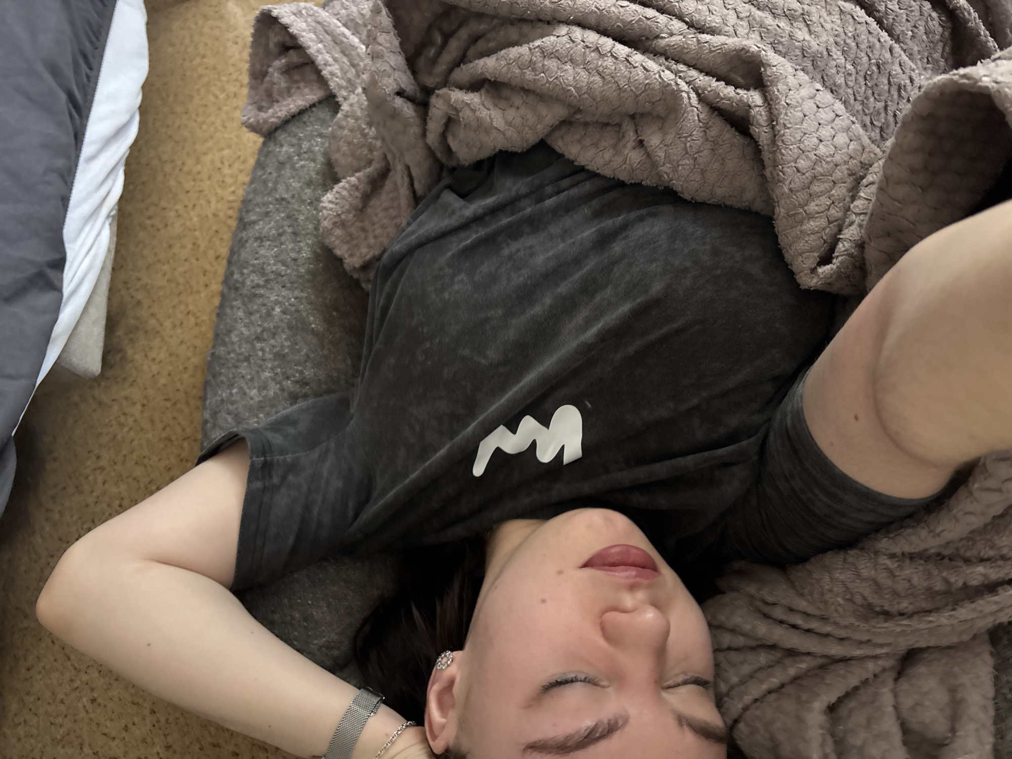 The scene depicts an indoor environment; the specific location within the city of Perm, Russia, cannot be determined from the visual evidence.
A single individual, identified as an adult female, is positioned lying on her left side, facing towards the lower-right of the frame. Her eyes are closed, indicating a state of rest or sleep. She possesses dark hair, a light skin tone, and is wearing dark lipstick. A small, shiny, floral or star-shaped earring is visible in her right ear. A silver-colored mesh watch with a rectangular face is secured on her right wrist, alongside a delicate silver-colored chain bracelet.
The individual is wearing a dark grey, short-sleeved t-shirt with a distinct white, stylized 'M' or 'W' shaped logo located on the left chest area. Her body is partially covered by a light brown or mauve-colored, textured blanket, exhibiting a patterned surface. Her head rests on a dark grey pillow or cushion. The visible floor surface is a light brown, low-pile carpet. In the upper portion of the frame, a dark grey bedspread or blanket is visible, layered over a white sheet, consistent with a bed.